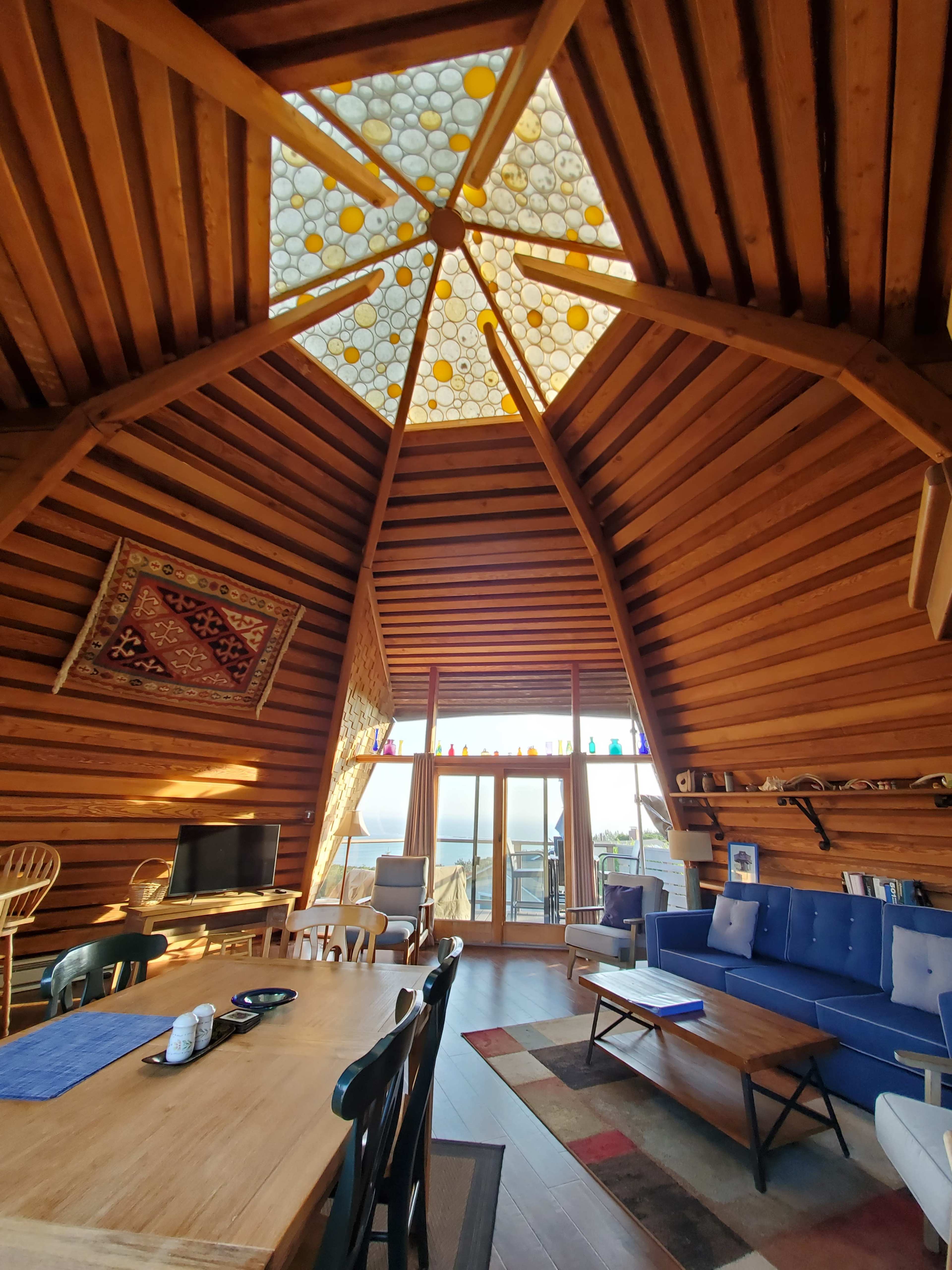 The interior of a wooden cabin features a high, angled ceiling with a glass skylight and a spacious living area furnished with a table, chairs, and a couch.