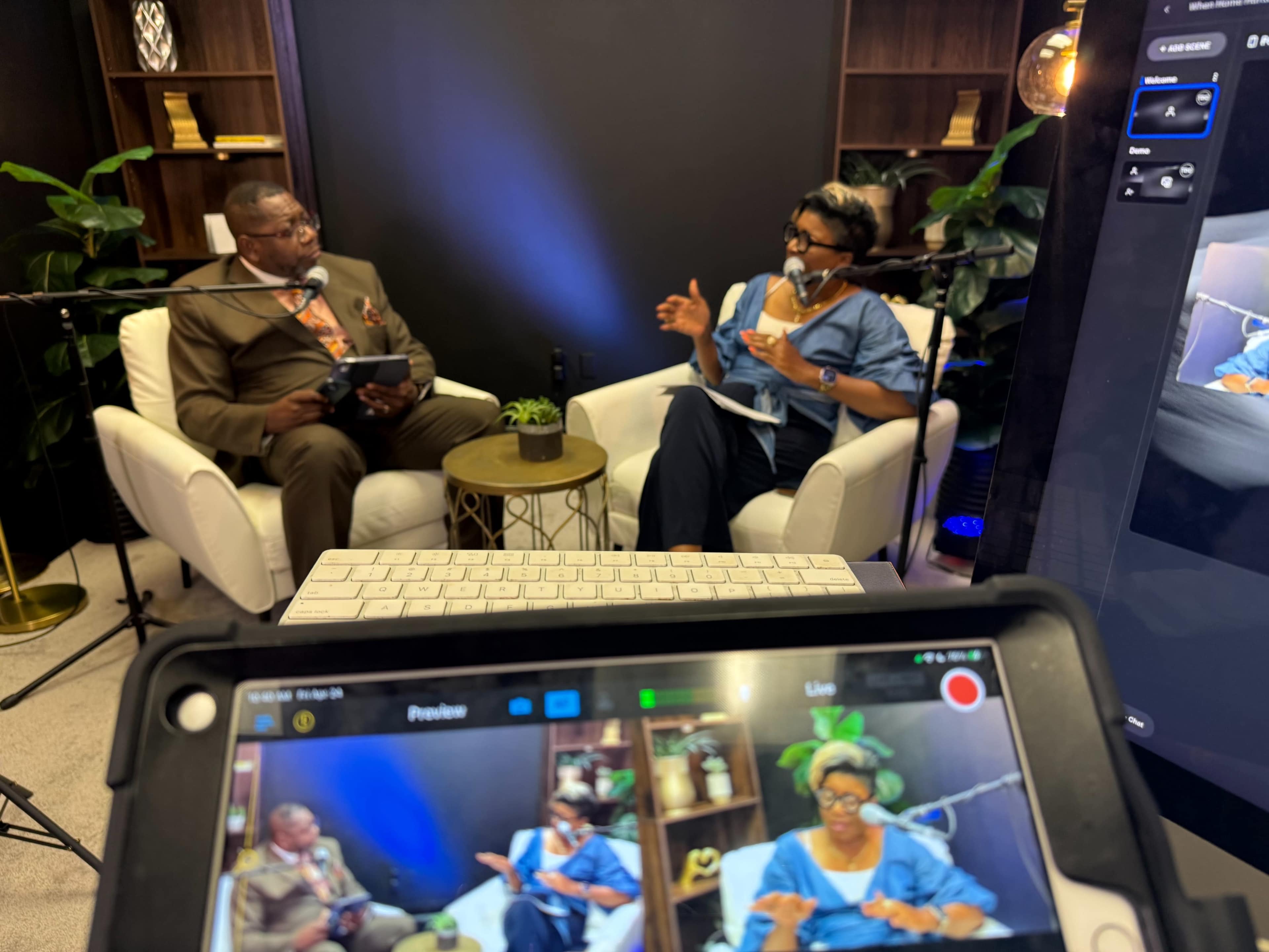 A man and a woman are engaged in conversation on a set with two white chairs, surrounded by plants and shelves, while a camera captures the scene.