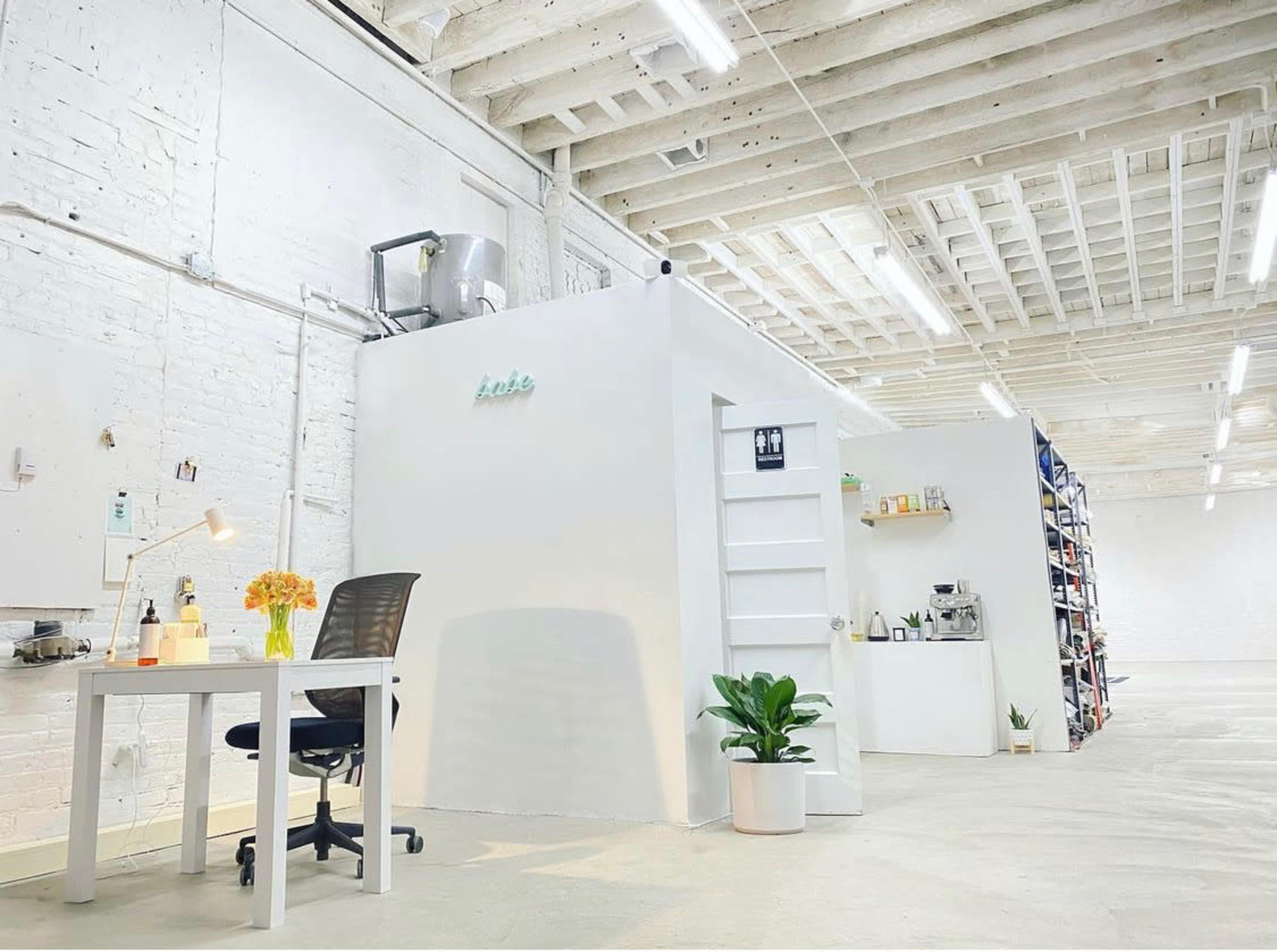A spacious, minimalist office environment with a white wall partition, a desk with a chair, a small plant, and shelves stocked with various items.