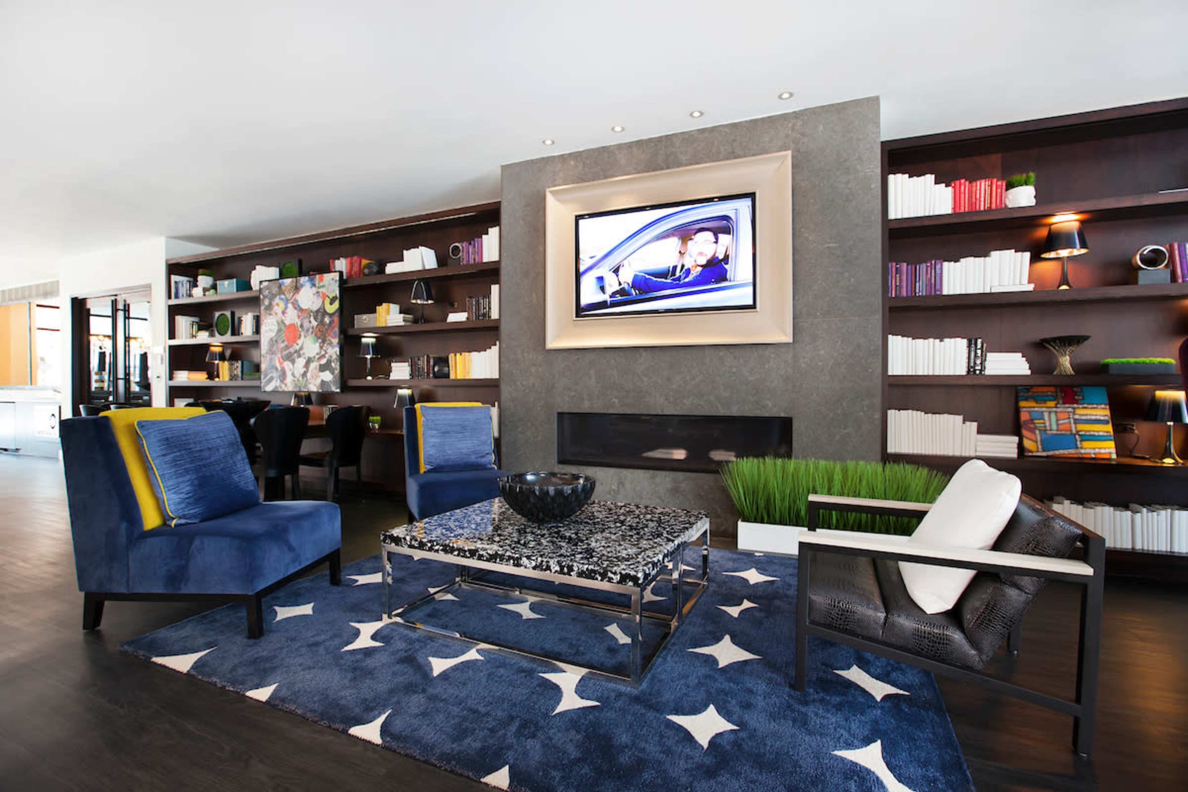 The image shows a modern lounge area featuring blue upholstered chairs, a patterned rug, a wooden coffee table, and a wall-mounted television above a sleek shelf with books and decor.