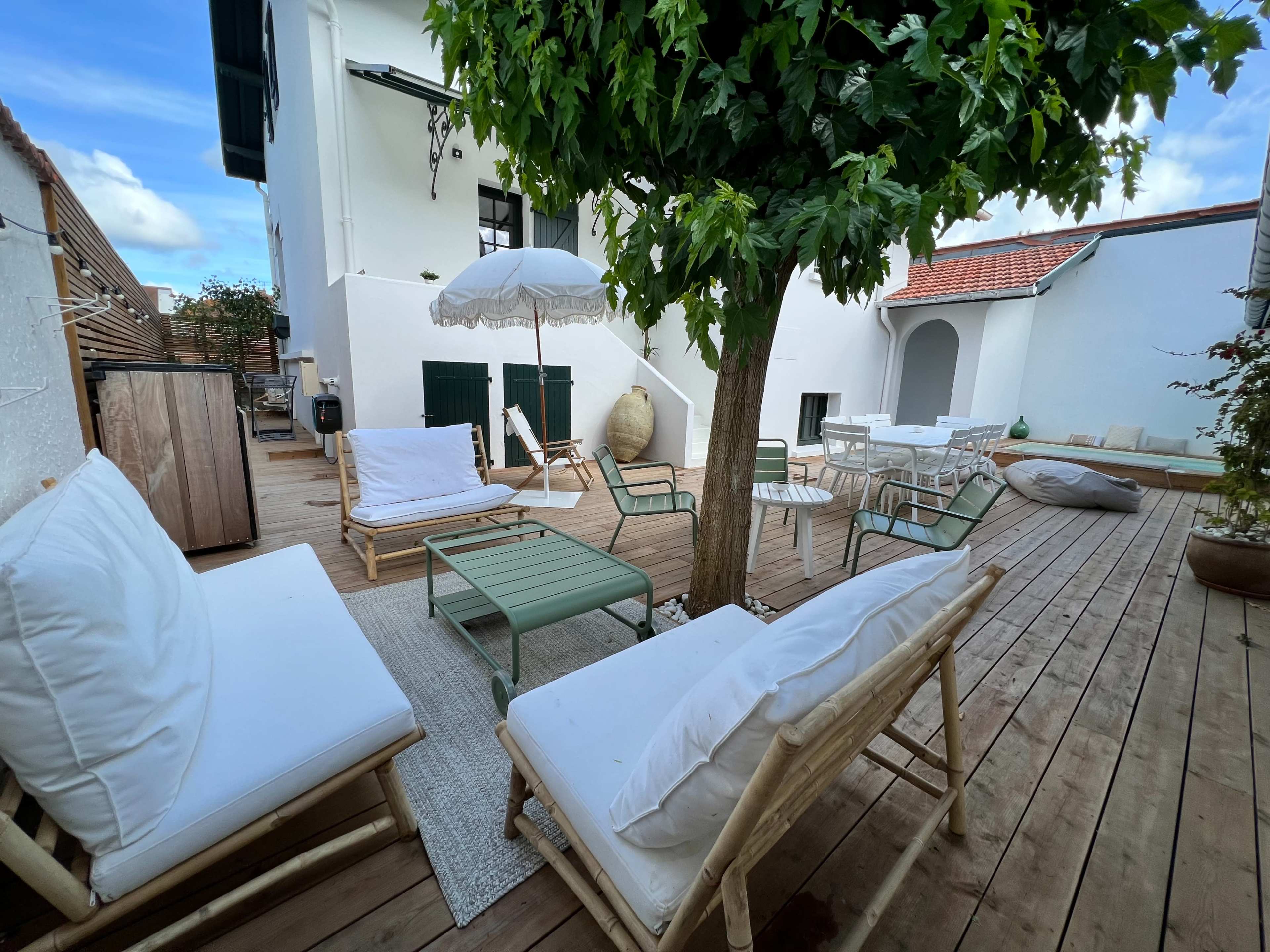 The image shows a wooden deck area with various seating arrangements, including lounge chairs and a table, surrounded by potted plants and a shaded umbrella.