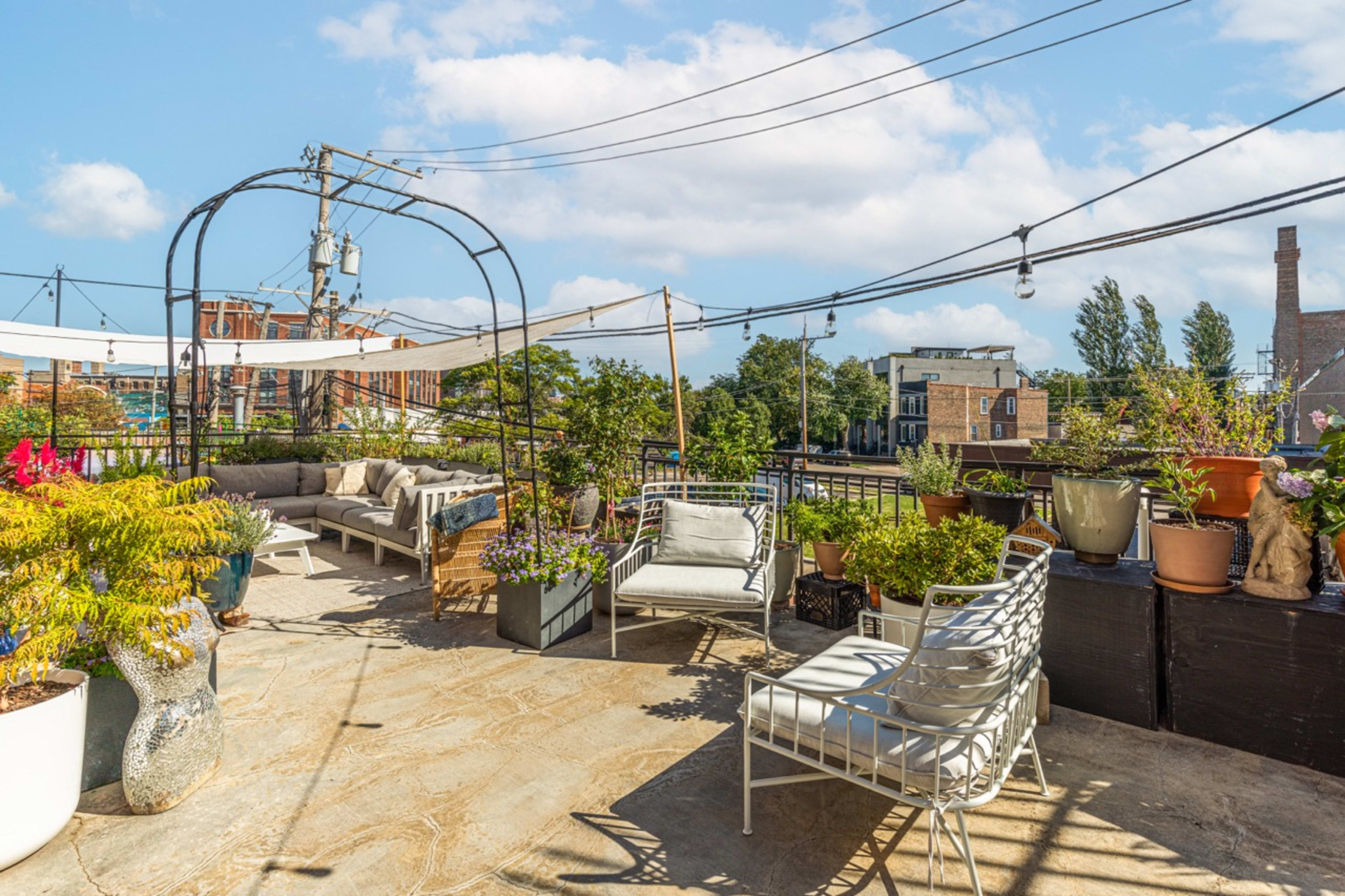 A rooftop terrace features a seating area with plants, white furniture, and overhead string lights against a backdrop of buildings and trees.