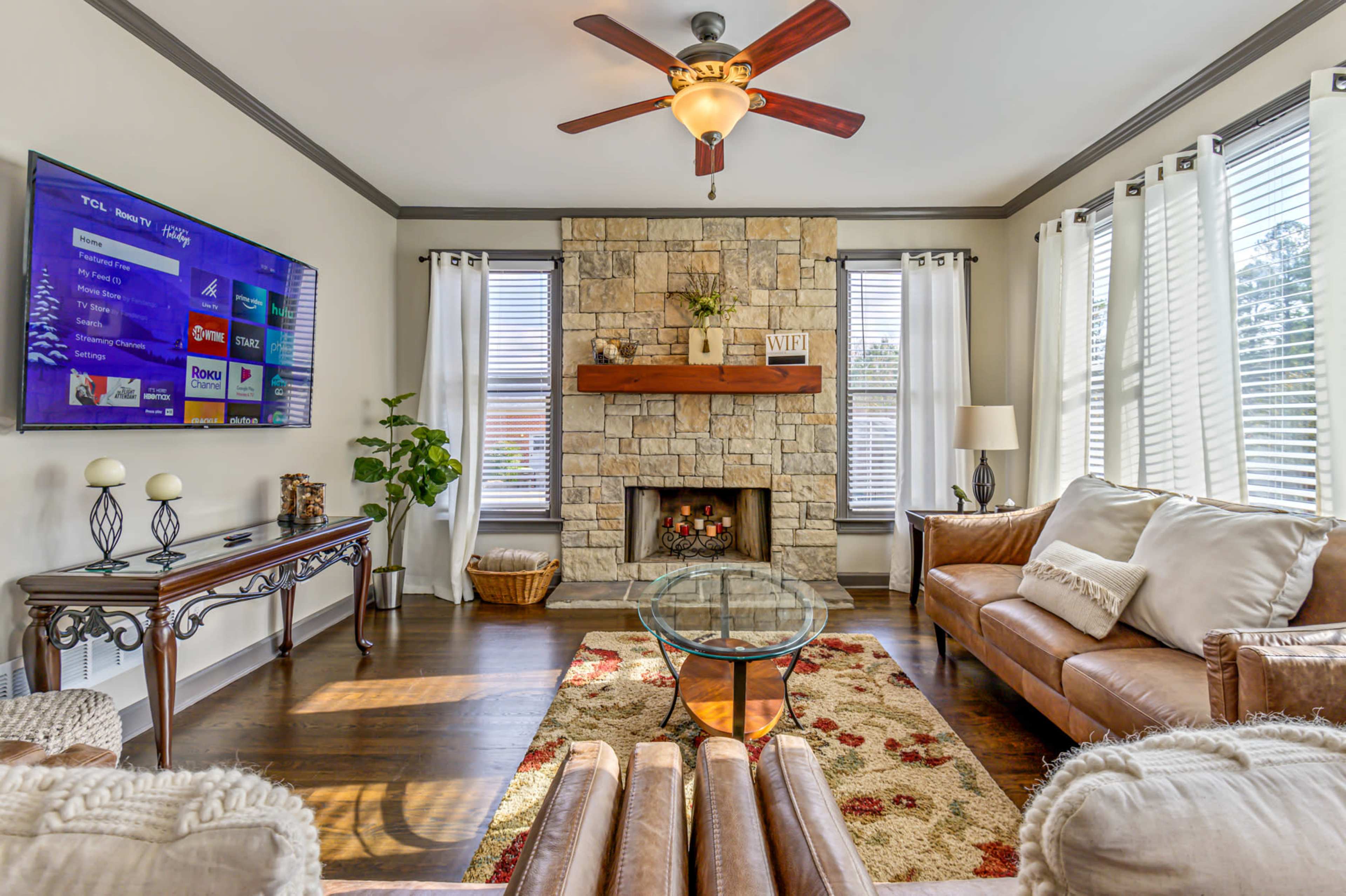 A cozy living room featuring a stone fireplace, a large television mounted on the wall, and comfortable seating arranged around a glass coffee table.