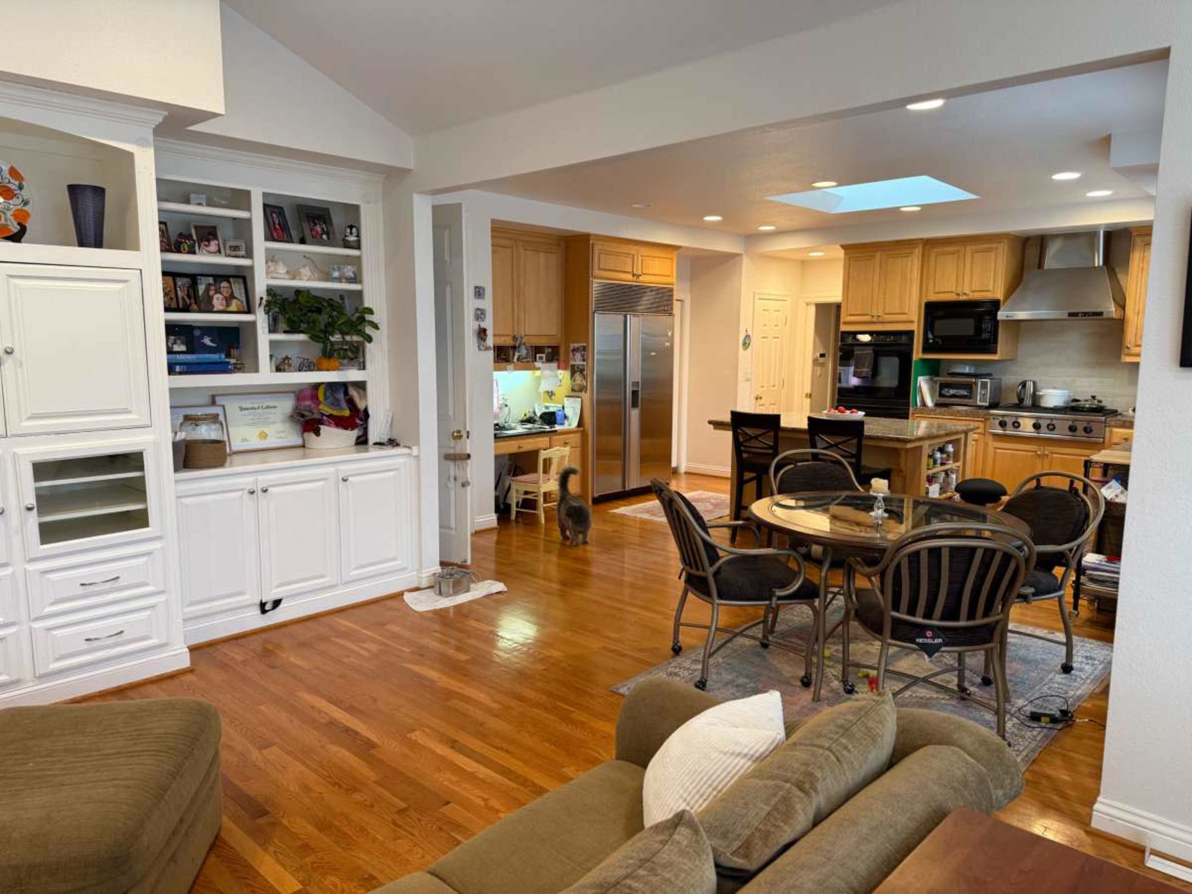 The image shows a spacious kitchen and dining area with wooden floors, a dining table, and a living room area featuring a couch and shelves.