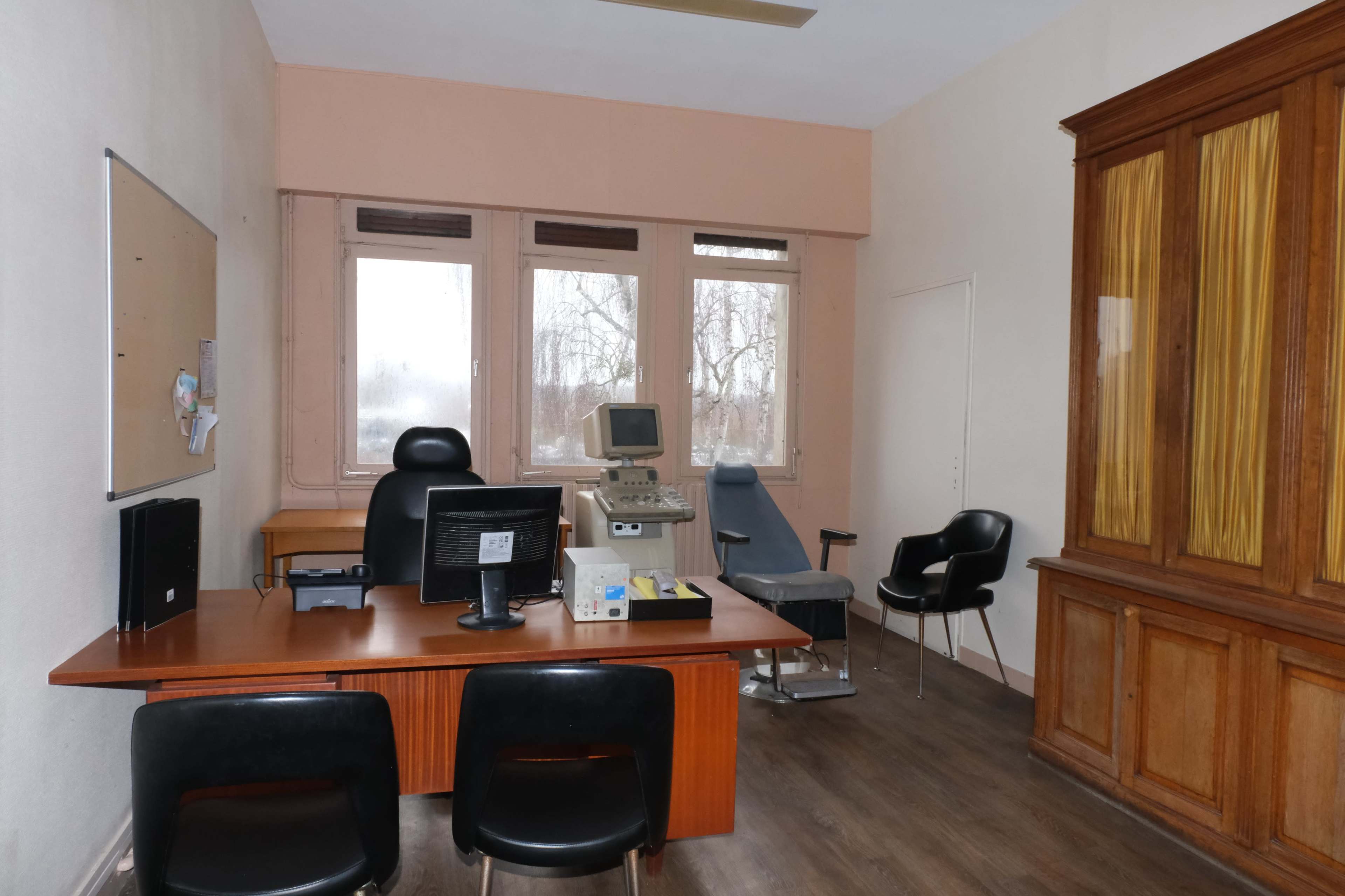 A small medical office featuring a desk with a computer, an examination chair, and a wooden cabinet.