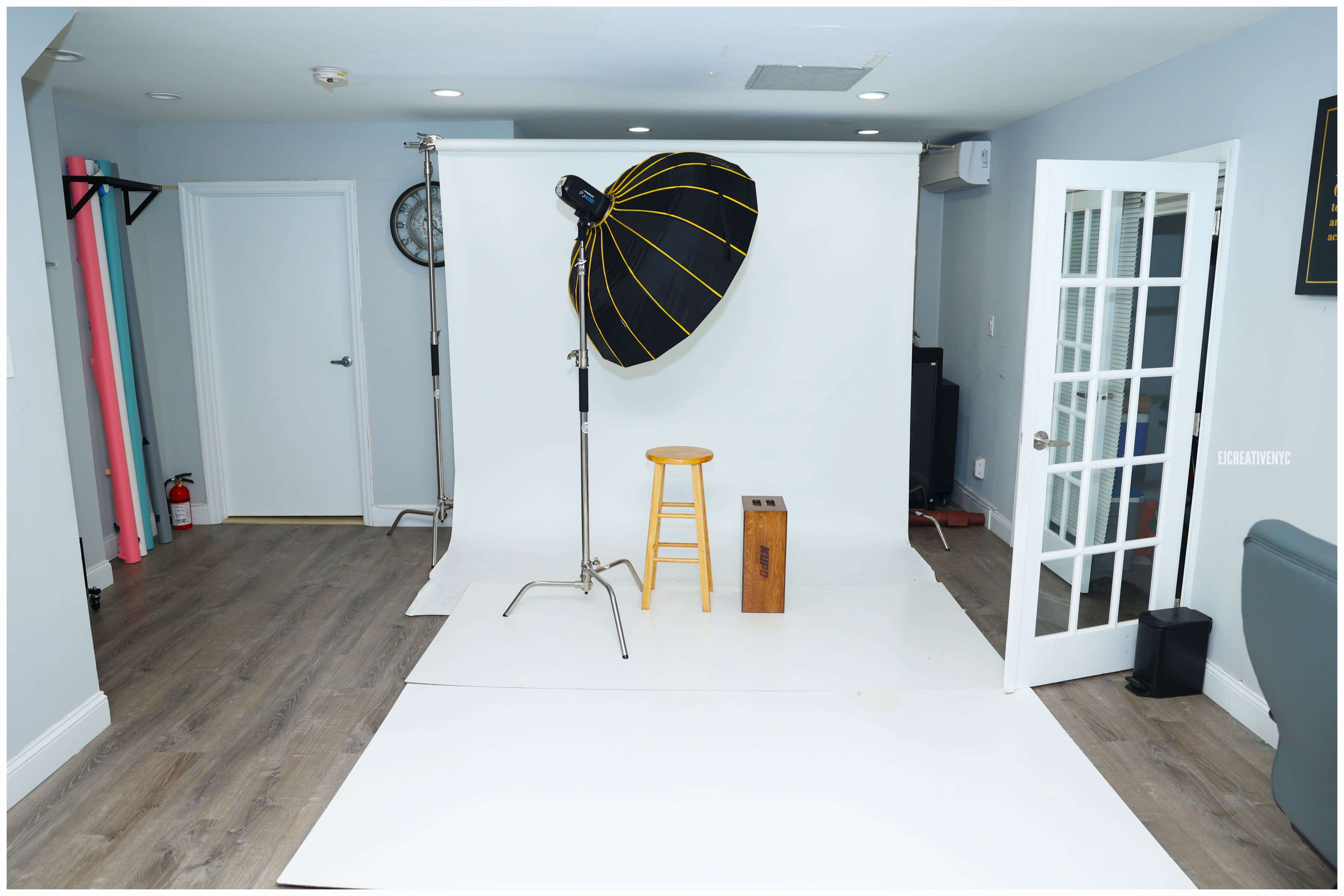 A photography studio with a white backdrop, a large softbox light, a wooden stool, and a small wooden box.