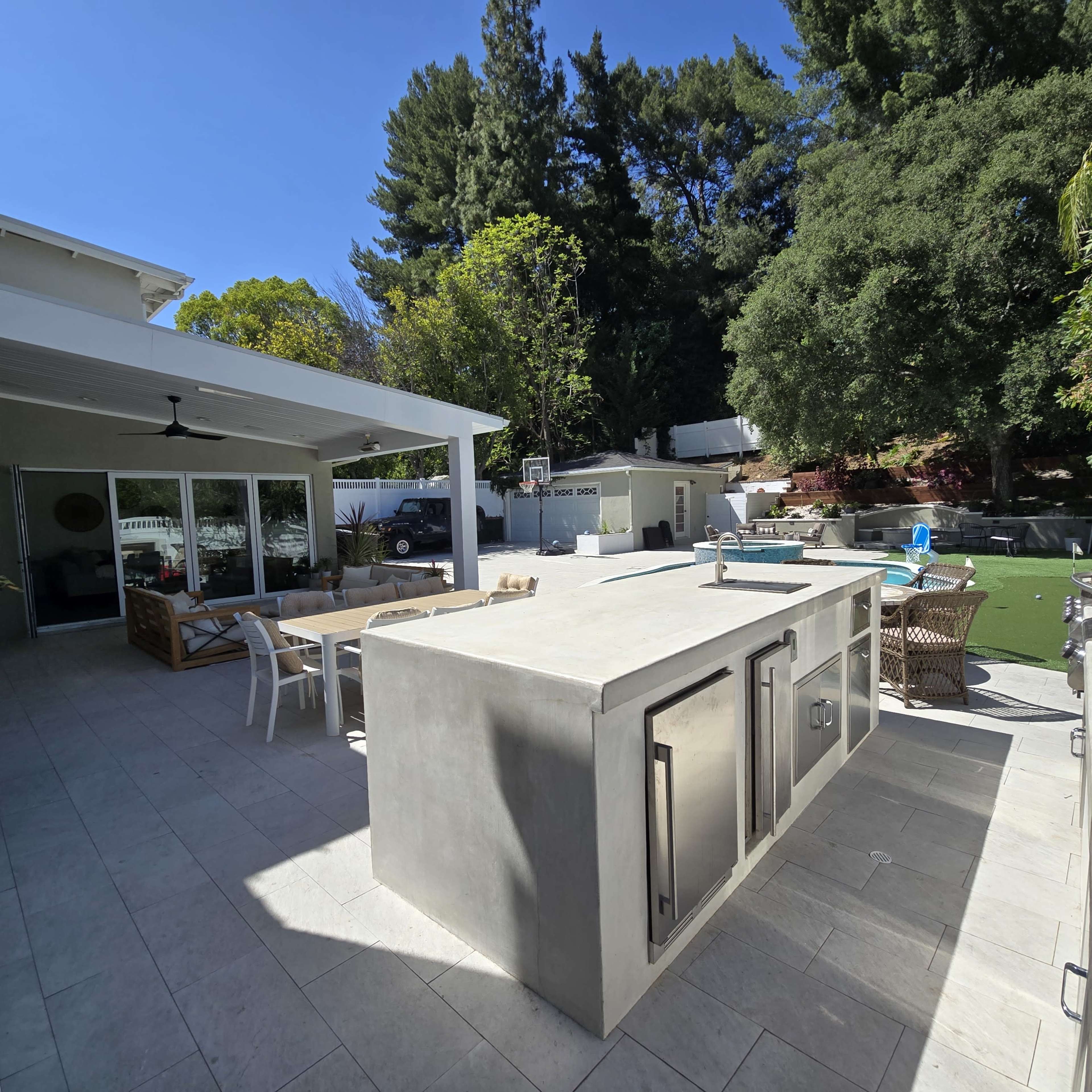The image features a modern outdoor kitchen with a concrete island, situated beside a dining area and surrounded by lush greenery and trees.