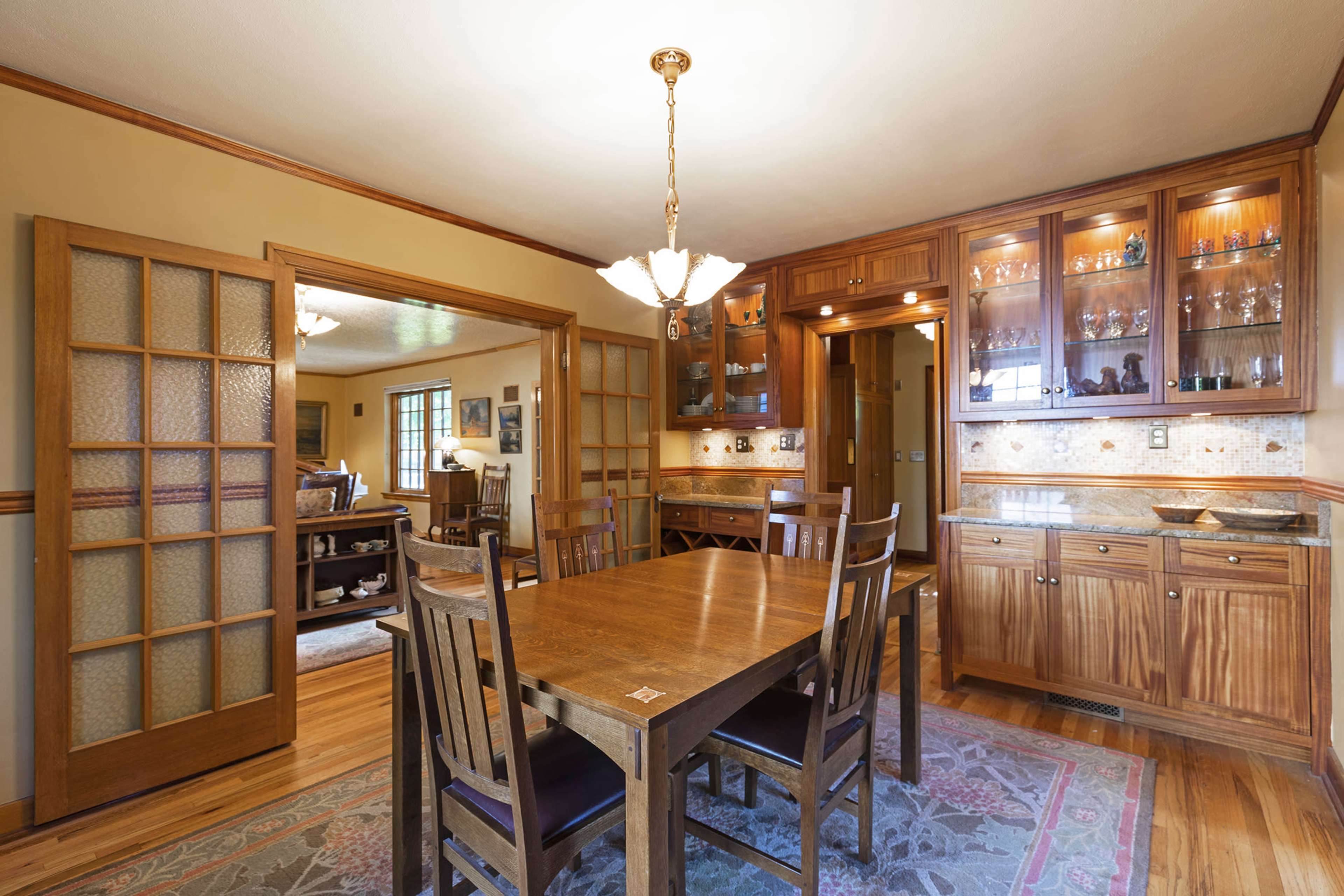 The image shows a wooden dining room with a large table and a built-in cabinet displaying glassware, featuring warm lighting and hardwood flooring.