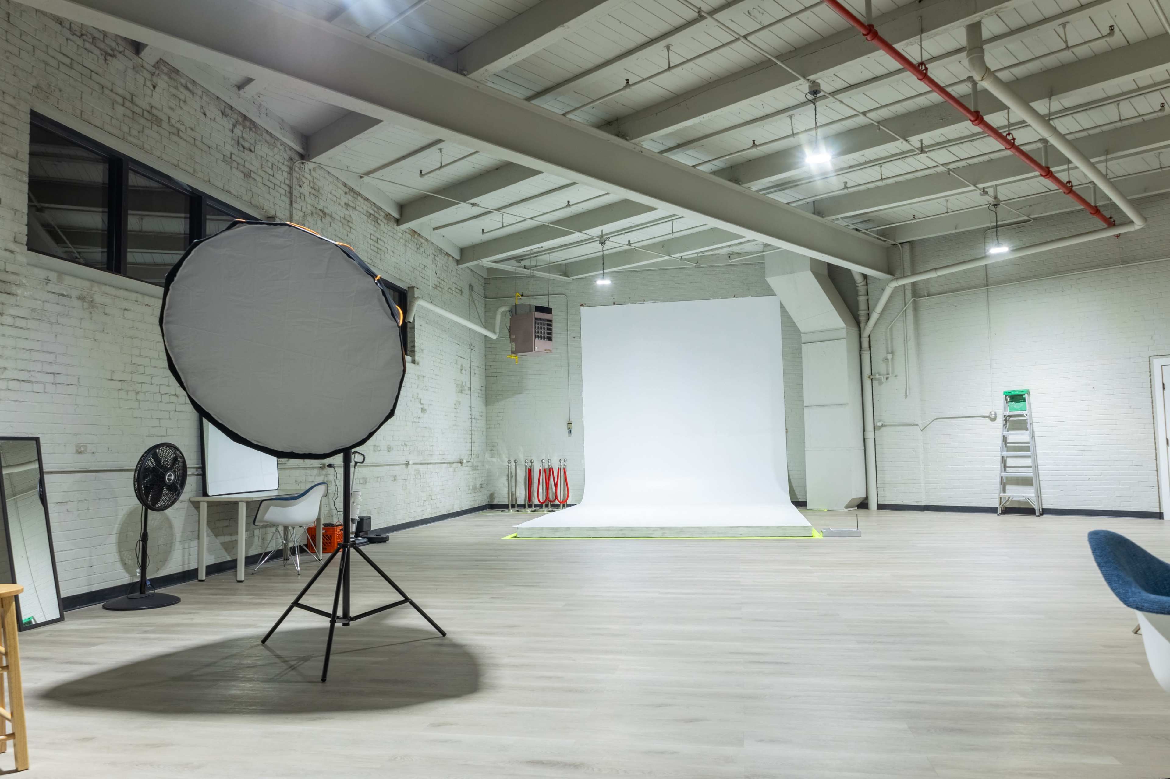 The image shows a well-lit photography studio with a white backdrop, a large round reflector on a stand, and minimal furnishings including a fan and a ladder.