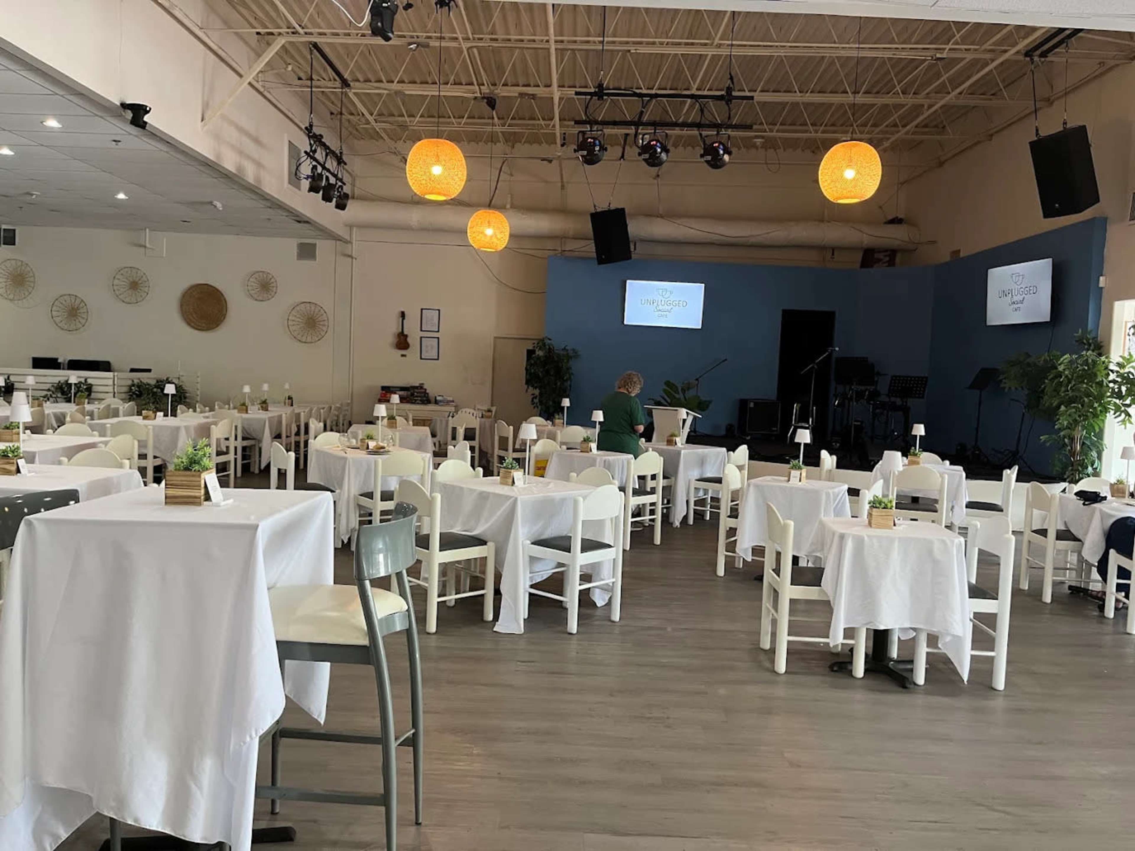 The image shows a spacious event venue with white tables covered in tablecloths, arranged with small centerpieces, and a small stage in the background.