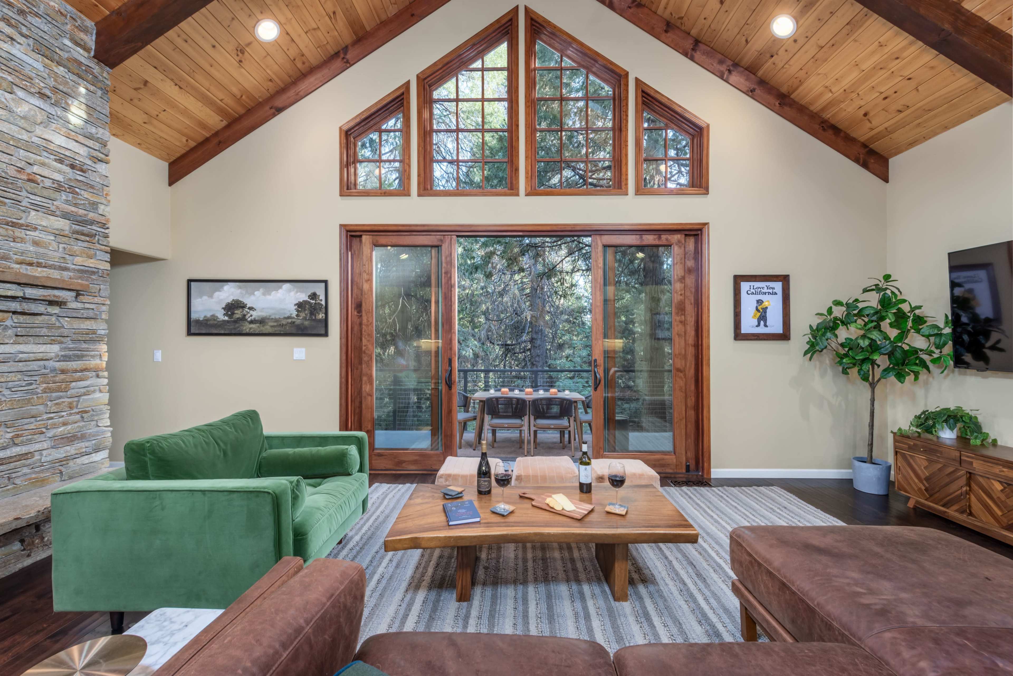 A cozy living room featuring a stone fireplace, green and brown couches, a wooden coffee table, and large windows with a view of greenery outside.