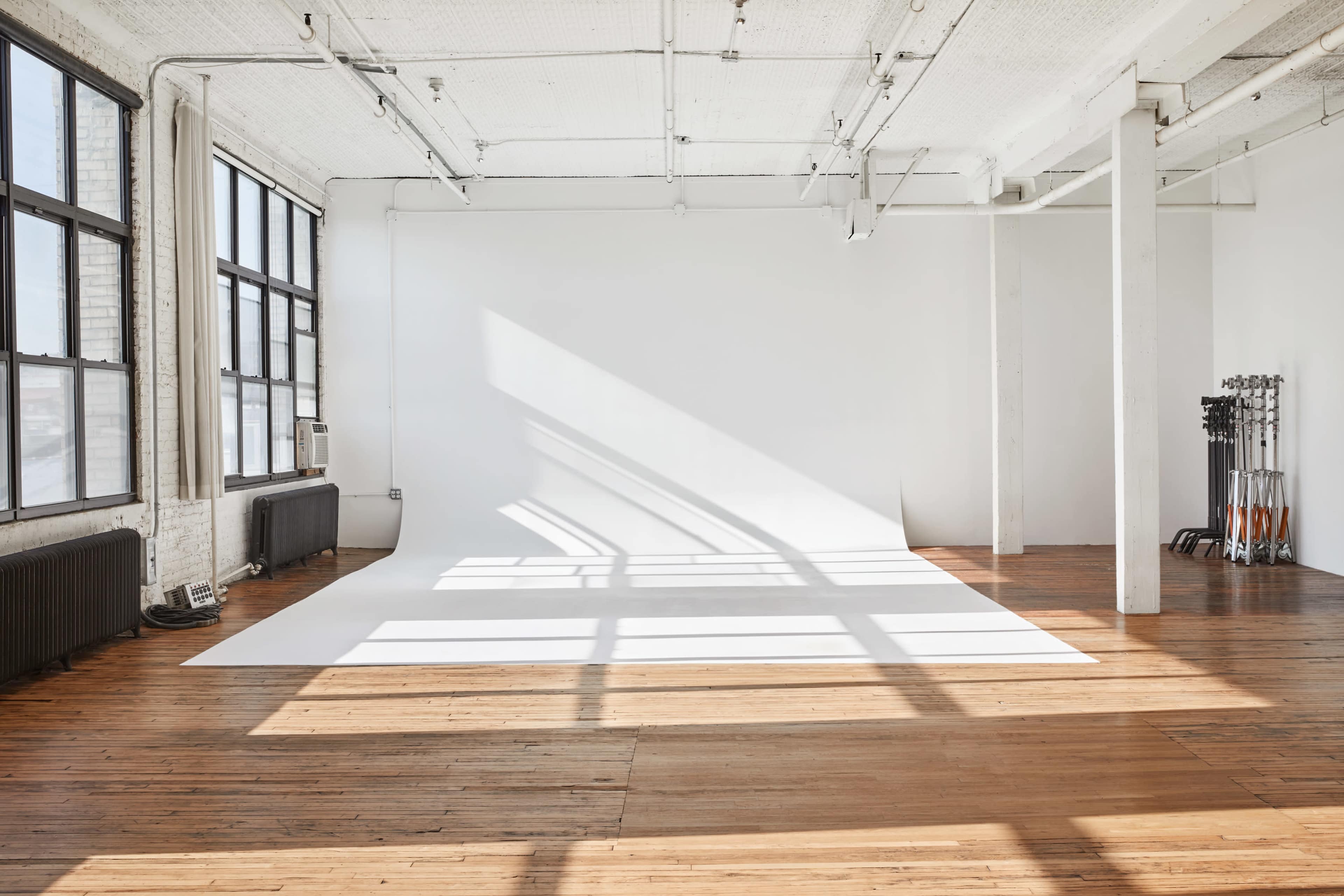A spacious, well-lit studio features a white backdrop and wooden flooring, with large windows casting shadows across the room.