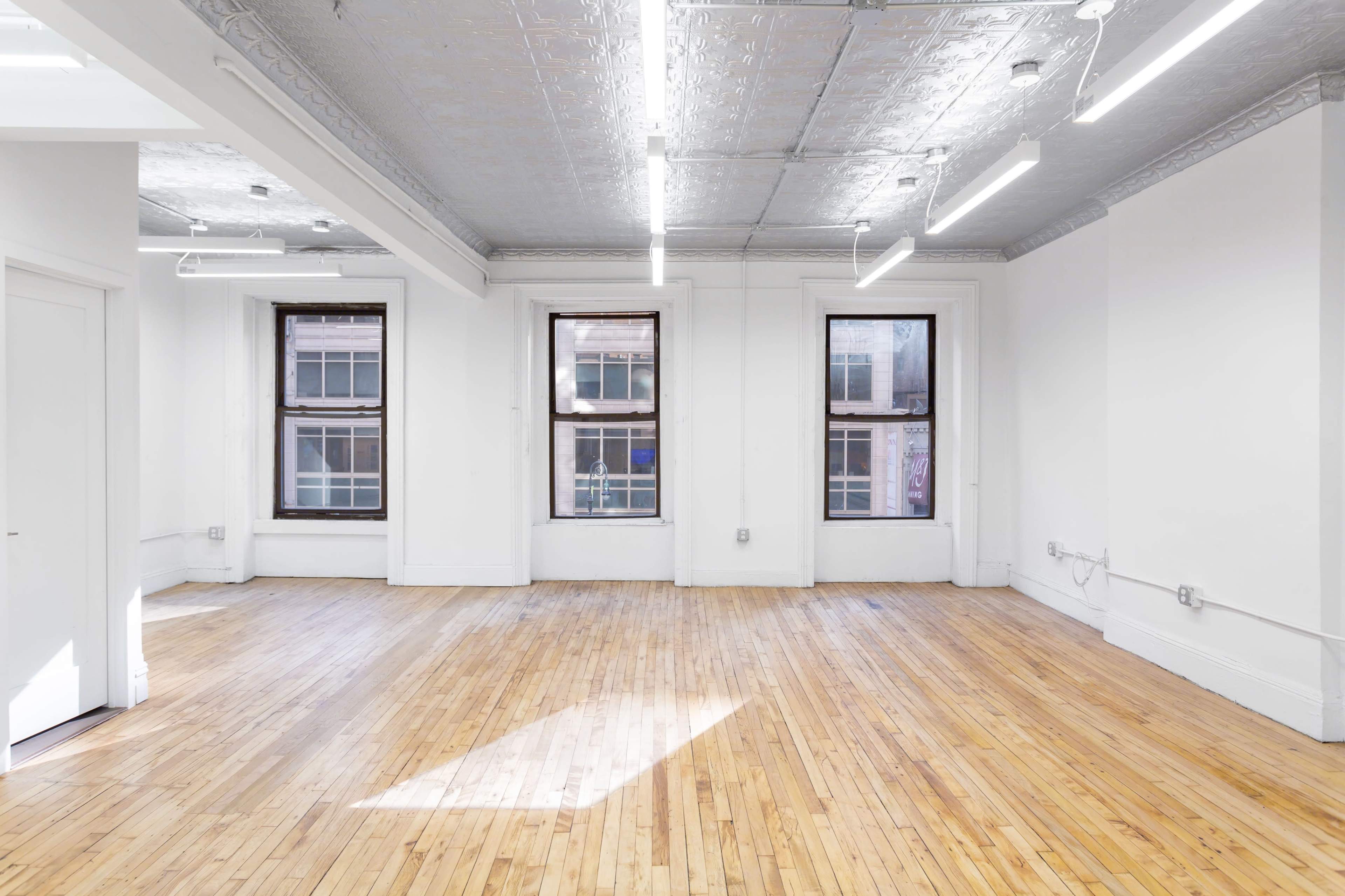 The image shows an empty, well-lit room with hardwood flooring and three large windows.