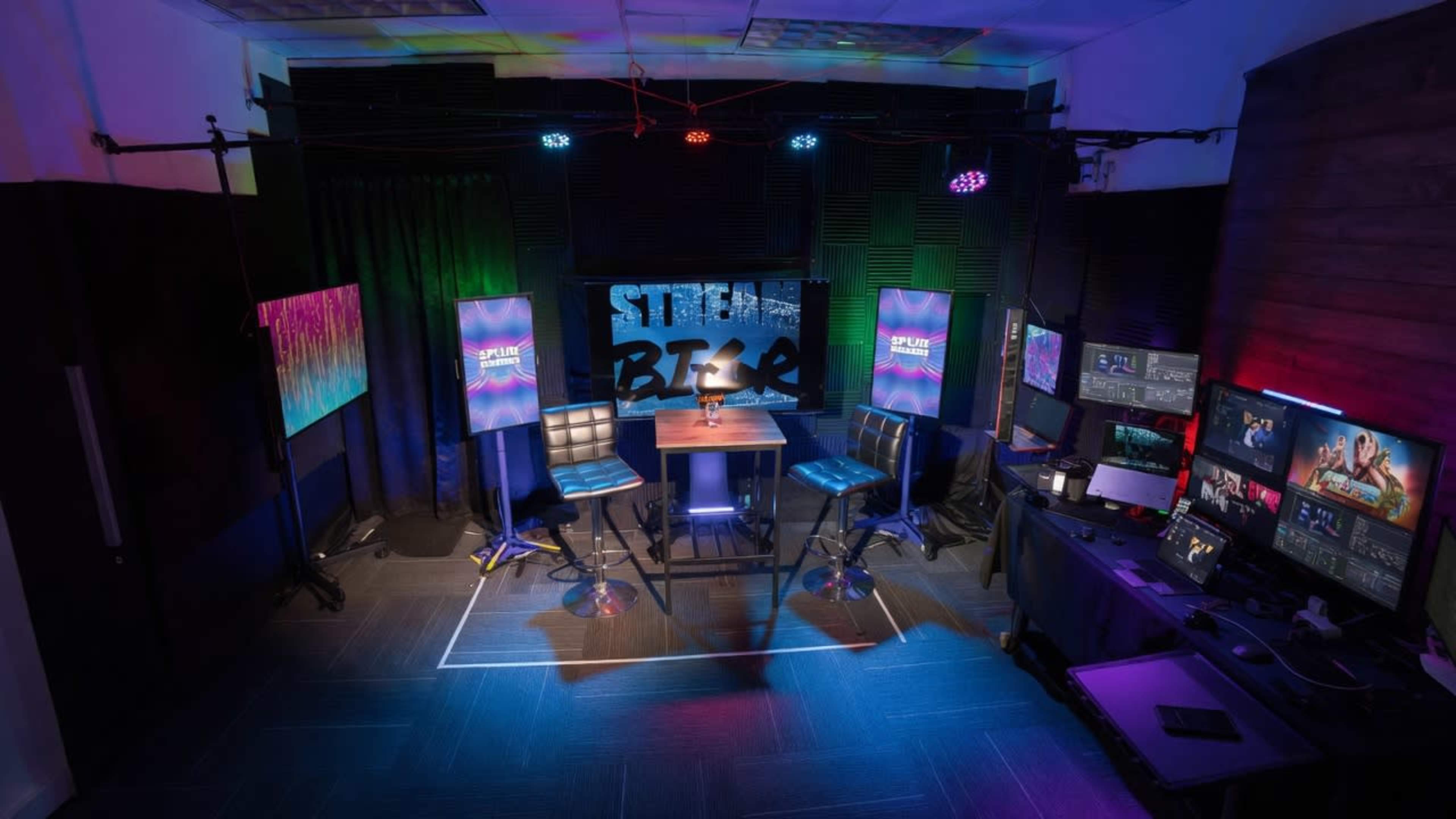 The image shows a well-equipped streaming studio featuring two high-top stools at a central table, surrounded by multiple monitors, colorful lighting, and screens displaying graphics.
