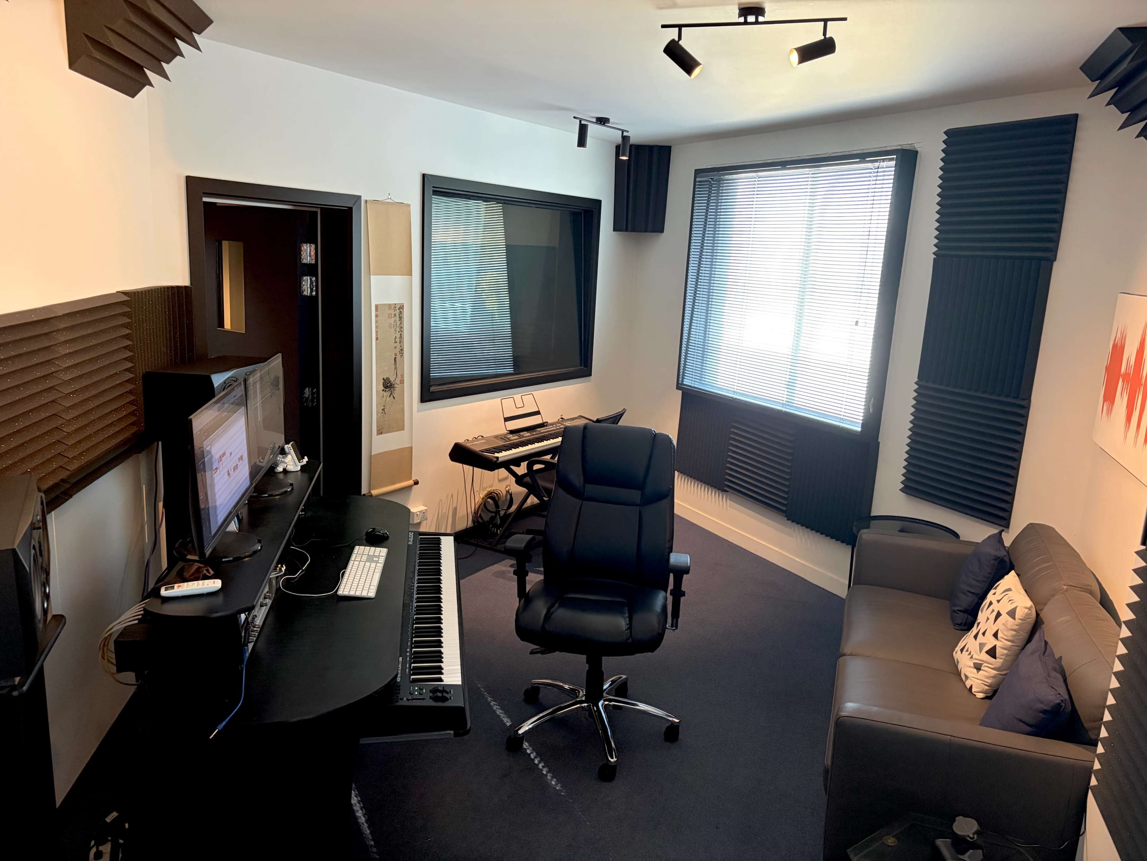 The image shows a small sound studio with acoustic panels, a keyboard, a computer setup on a black desk, and a gray couch.