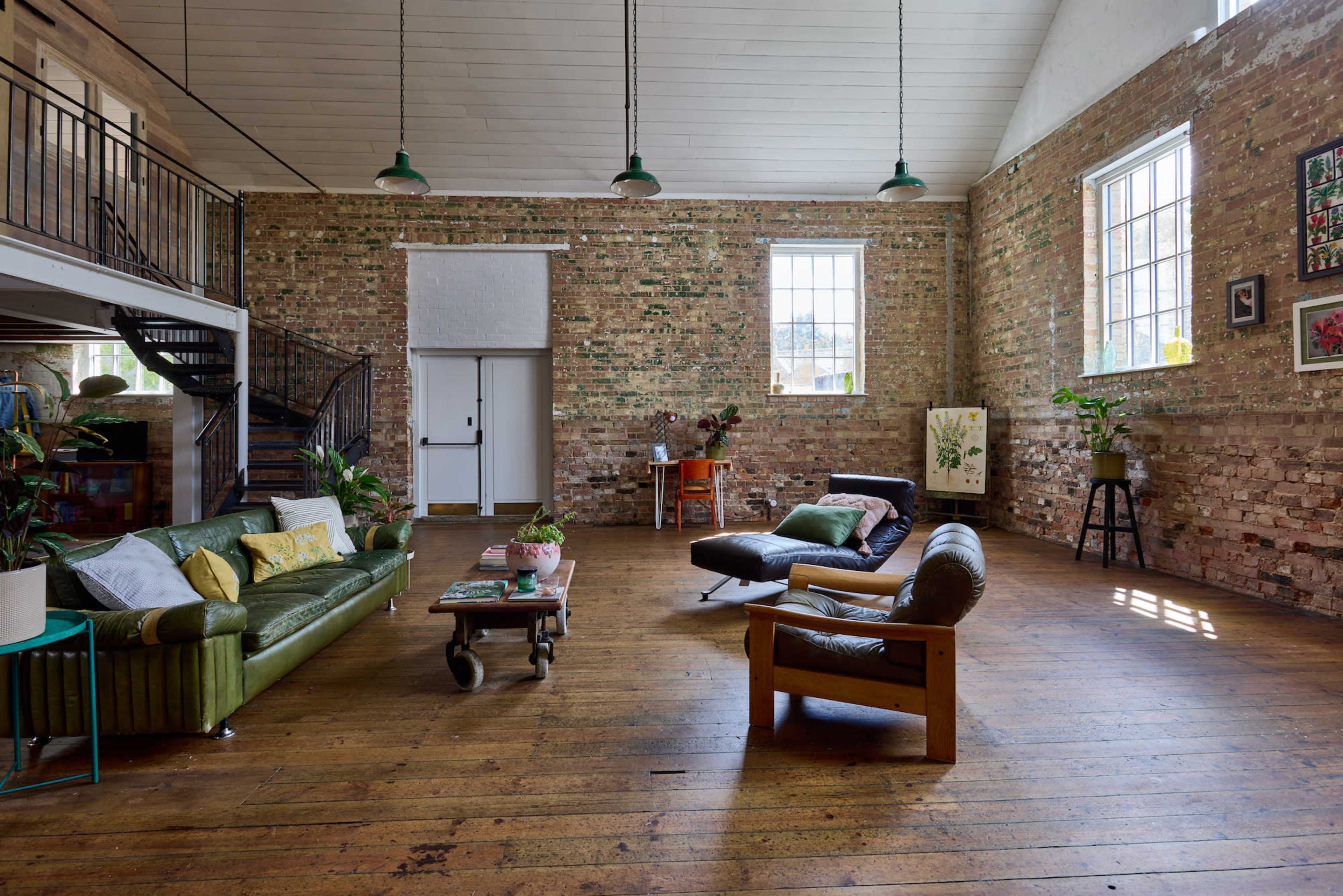 The spacious room features exposed brick walls, large windows, and a mix of modern and vintage furniture, including green and brown sofas and wooden flooring.