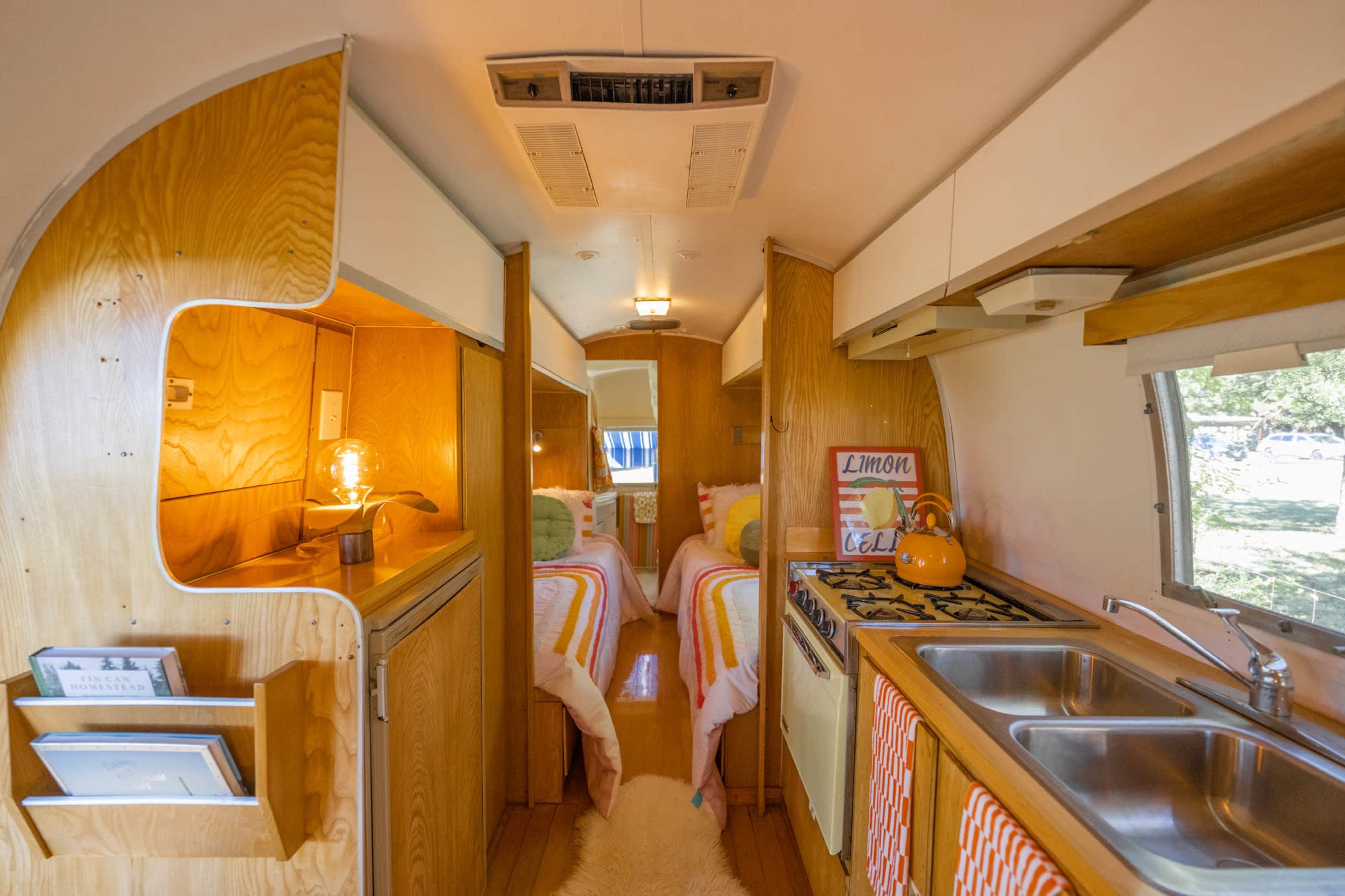 The image shows the interior of a vintage camper trailer with two beds, a kitchenette, and wooden paneling.