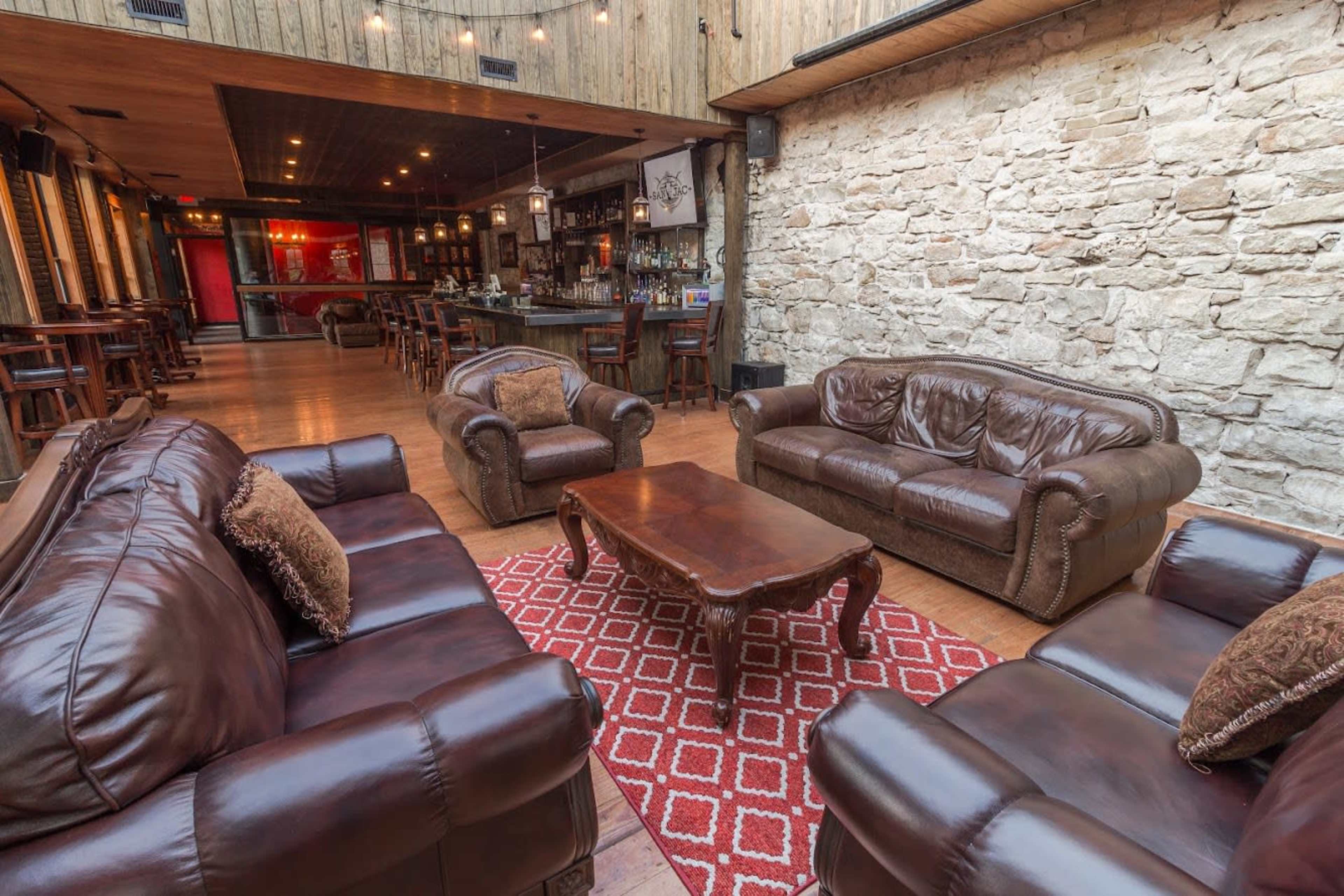 A seating area features brown leather sofas arranged around a wooden coffee table on a patterned rug, with a stone wall and bar in the background.