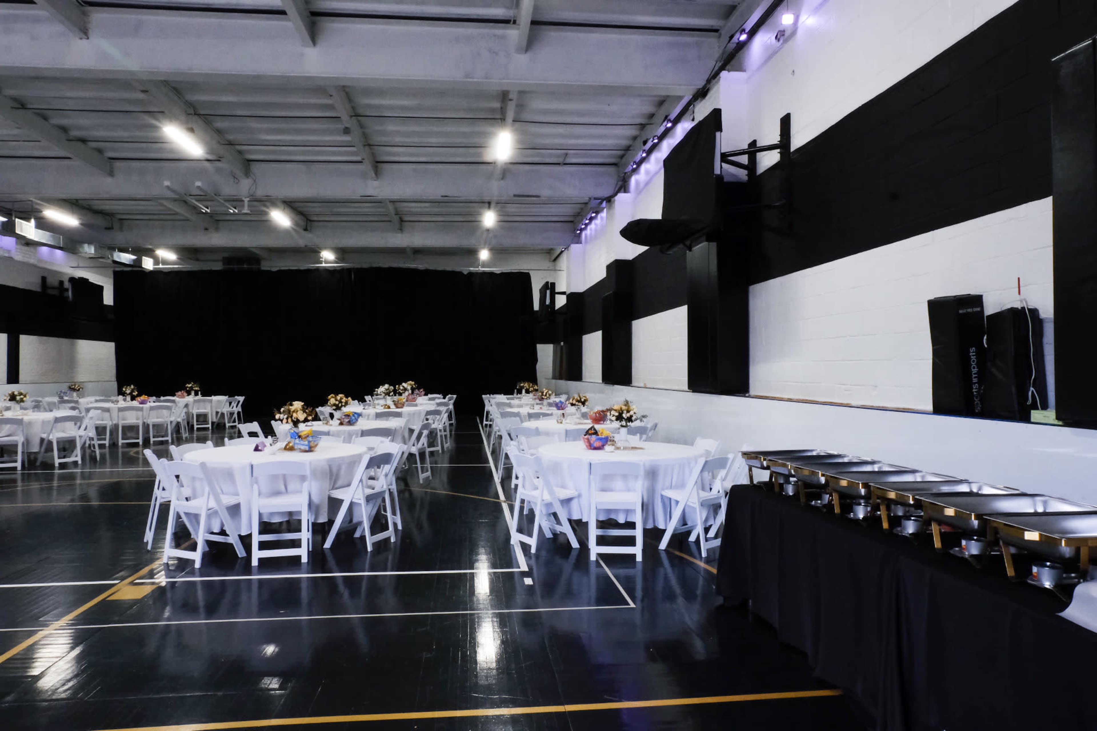 The image shows a large indoor space set up for an event, featuring round tables with white tablecloths arranged in rows and a buffet table along one wall.