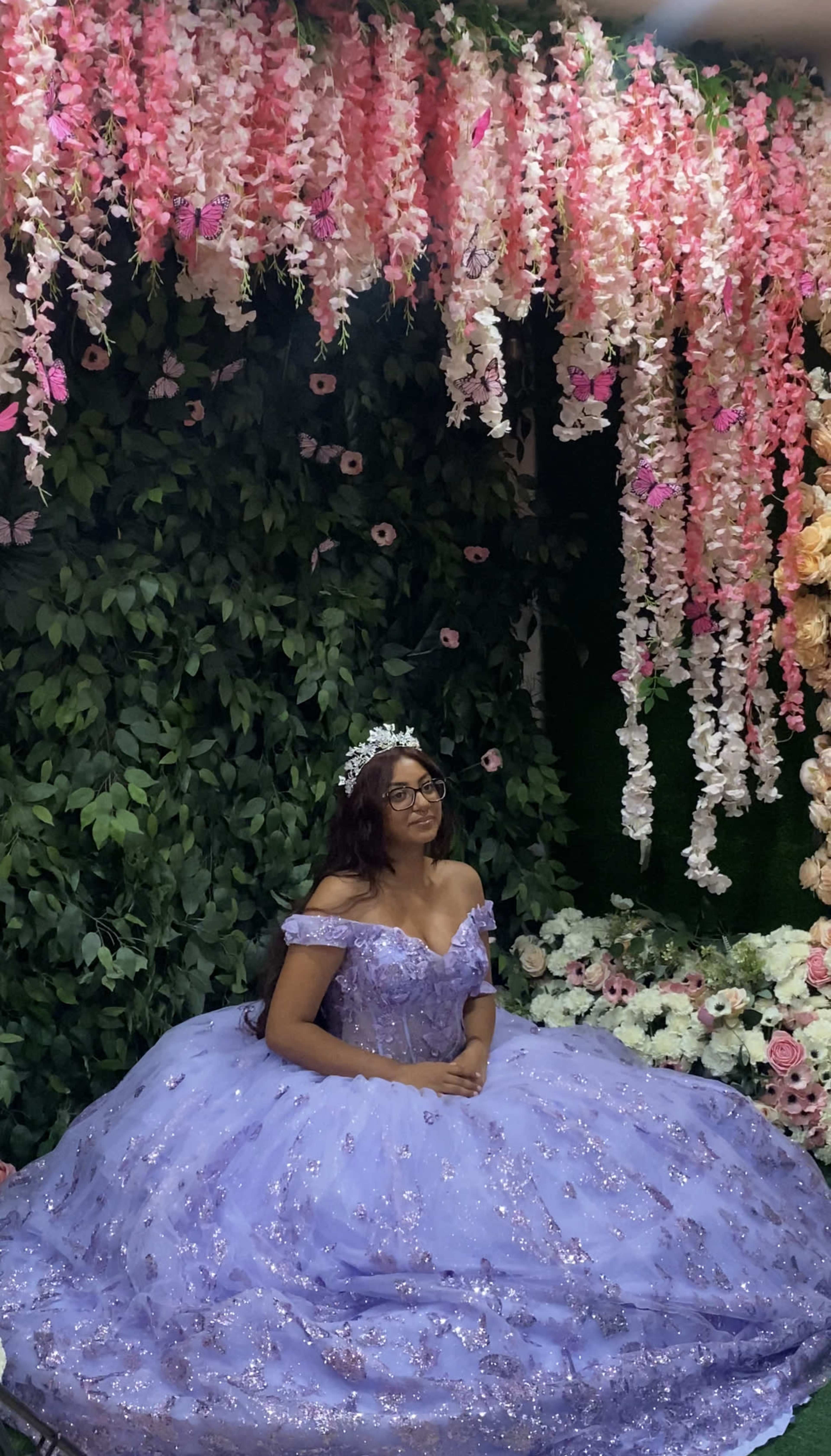 A person in a lavender gown sits in front of a floral backdrop adorned with hanging pink and white decorations.