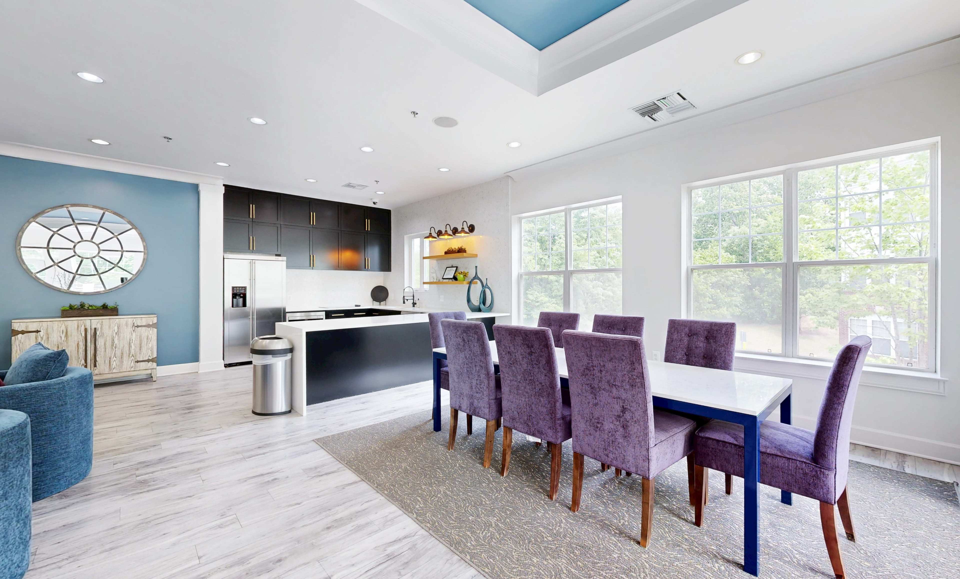 The image shows a modern dining area with a large table surrounded by purple chairs, adjacent to a sleek kitchen with dark cabinetry and a spacious layout.