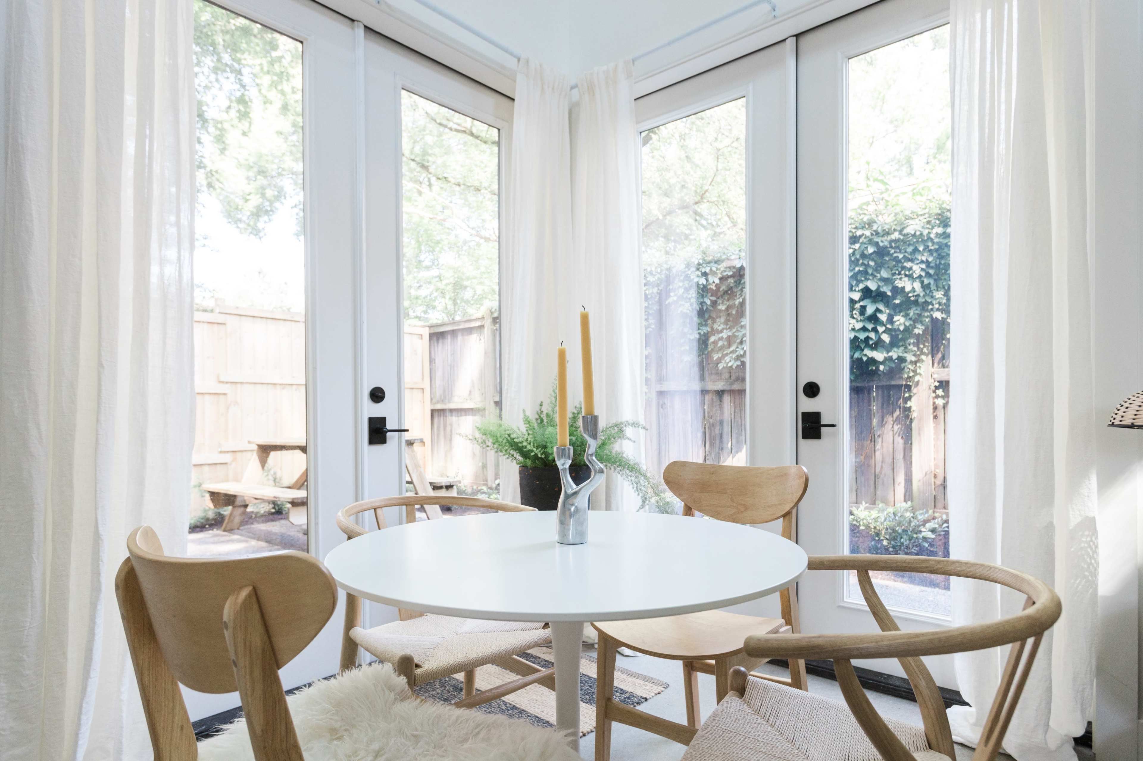 A round white table with two wooden chairs is set near glass doors that open to a garden, with light-colored curtains framing the view.