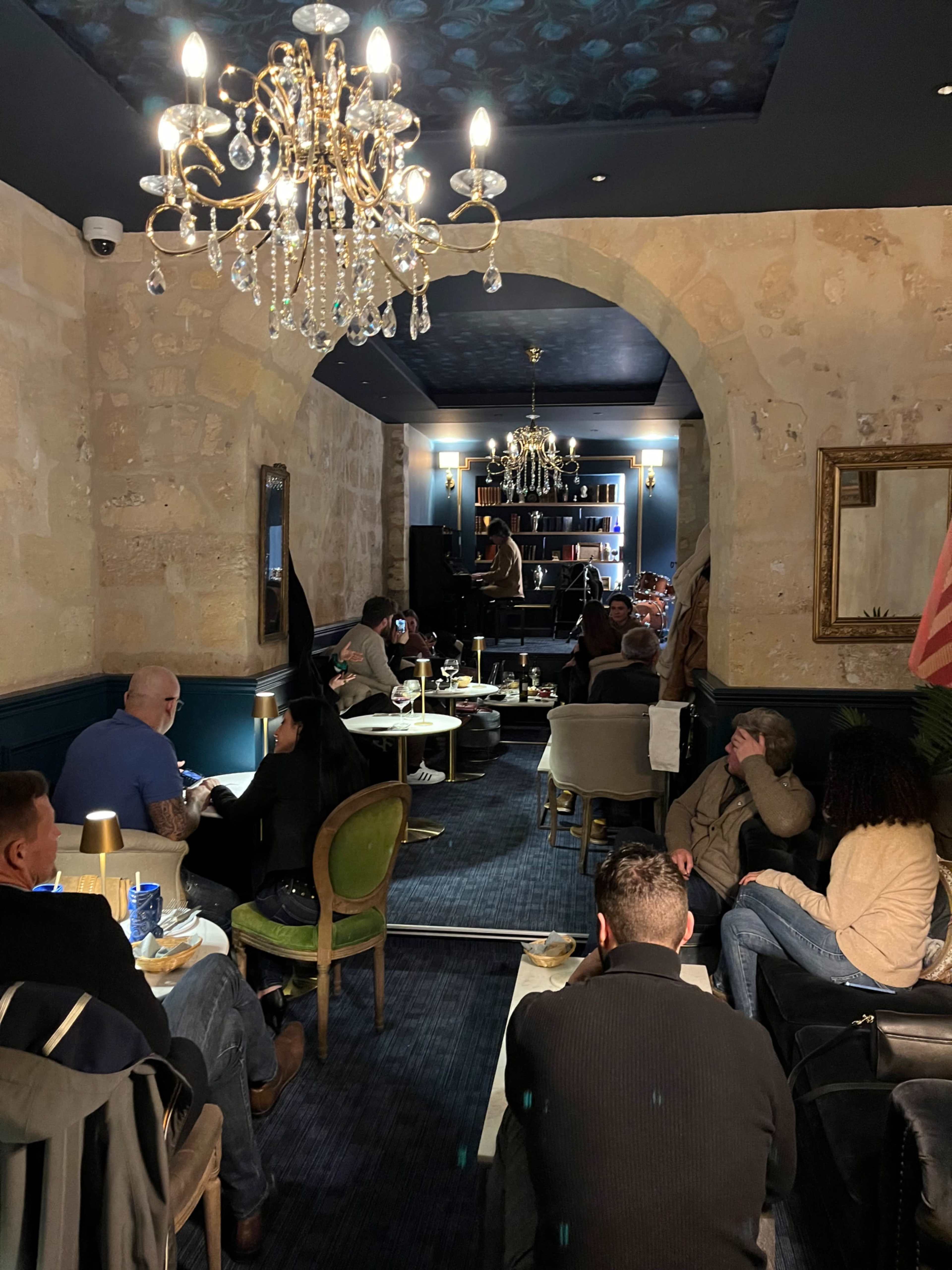 The image shows a dimly lit restaurant with patrons seated at tables, some enjoying drinks while a musician plays piano in the background.