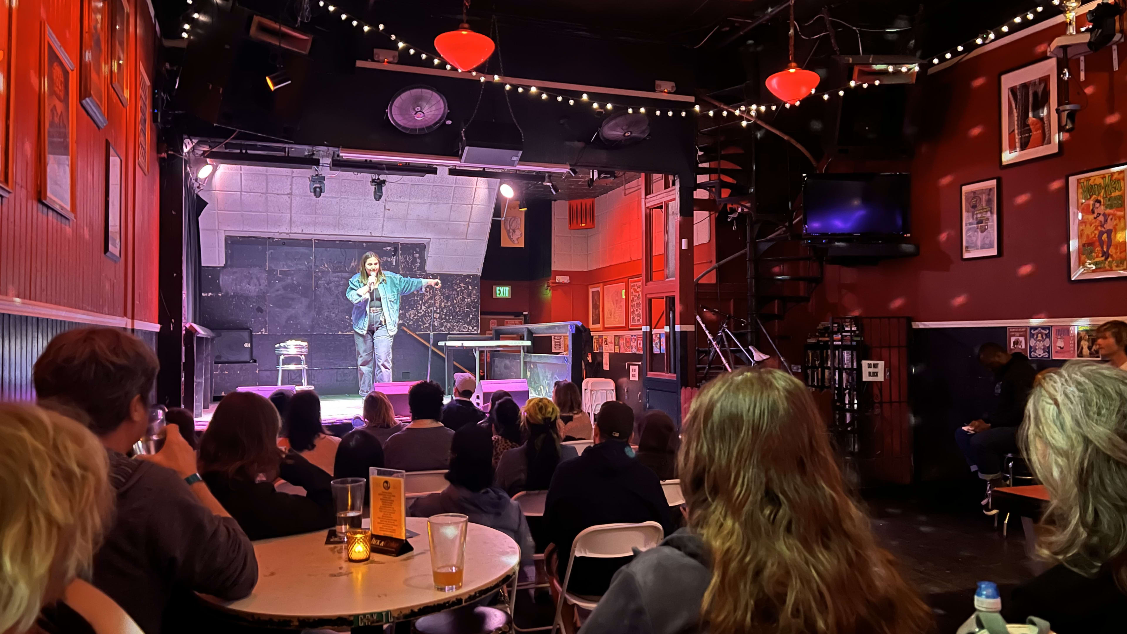 A stand-up comedian performs on stage in a dimly lit comedy club filled with seated audience members.