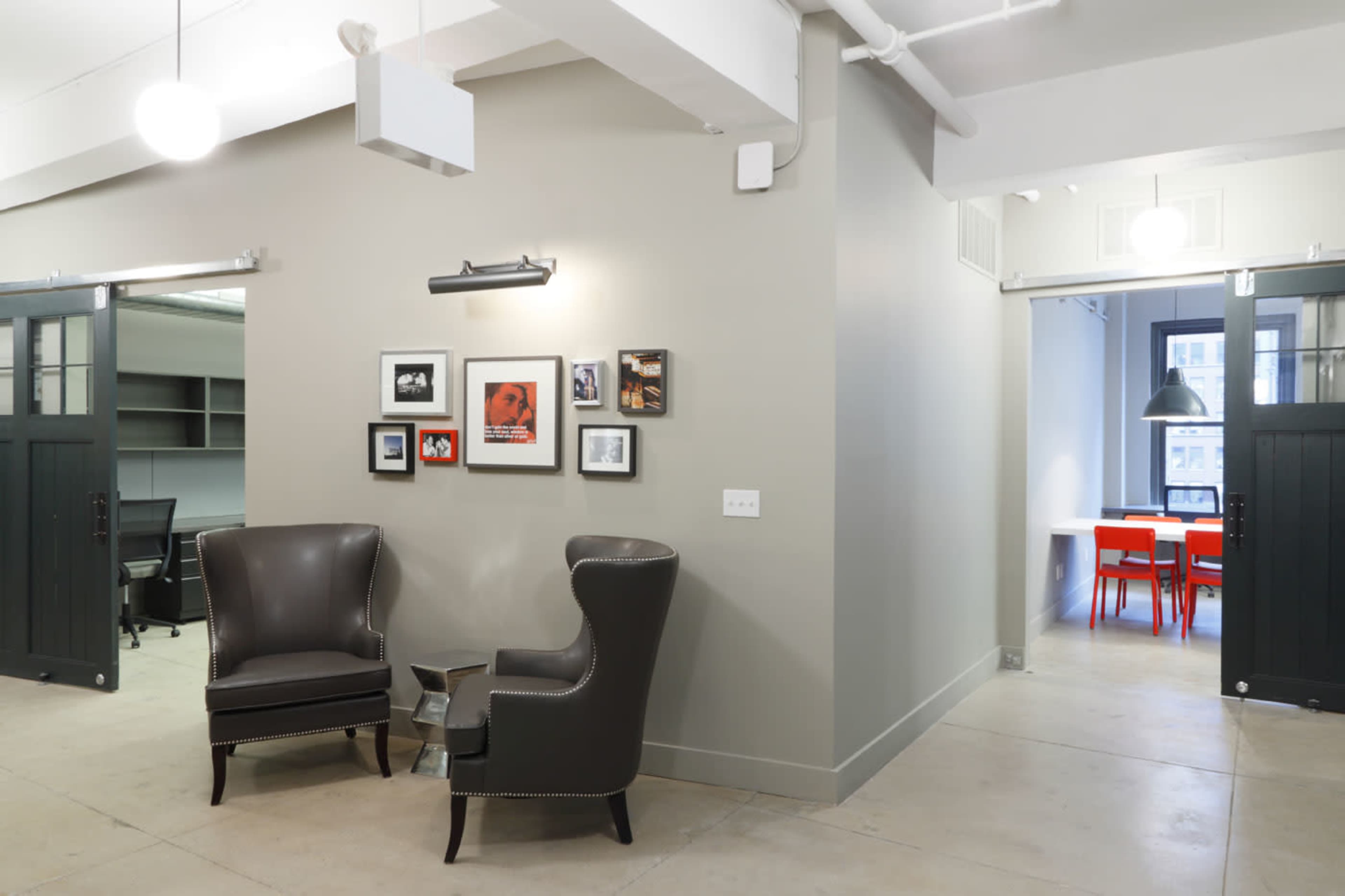 The image shows a modern office space with two dark leather chairs positioned near framed photos on a gray wall, leading to a conference area with red chairs in the background.