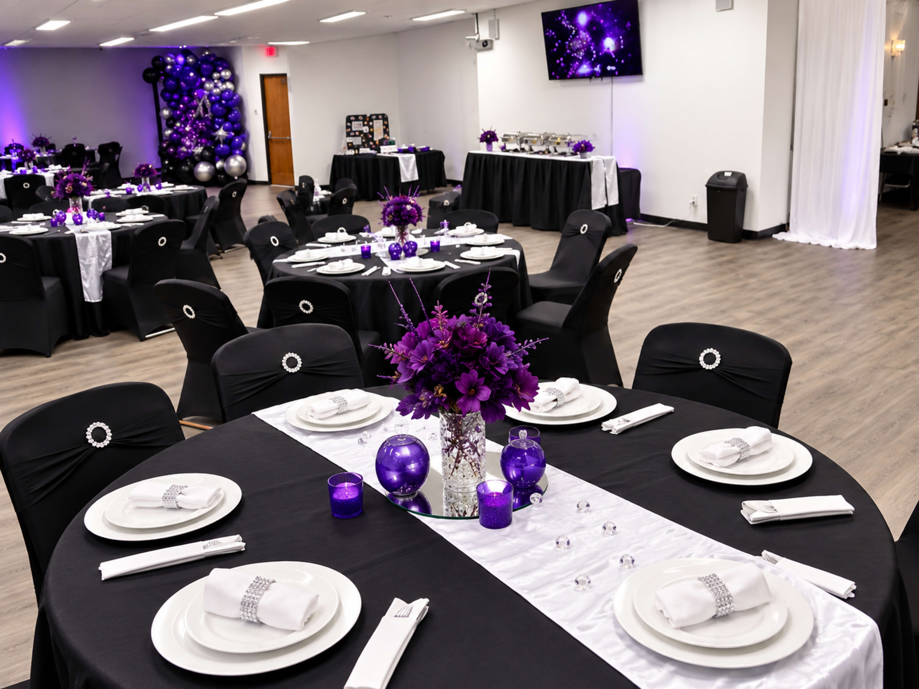 The image shows a banquet hall set for an event, featuring round tables adorned with black tablecloths, white runners, and purple floral centerpieces, with matching decor surrounding the room.