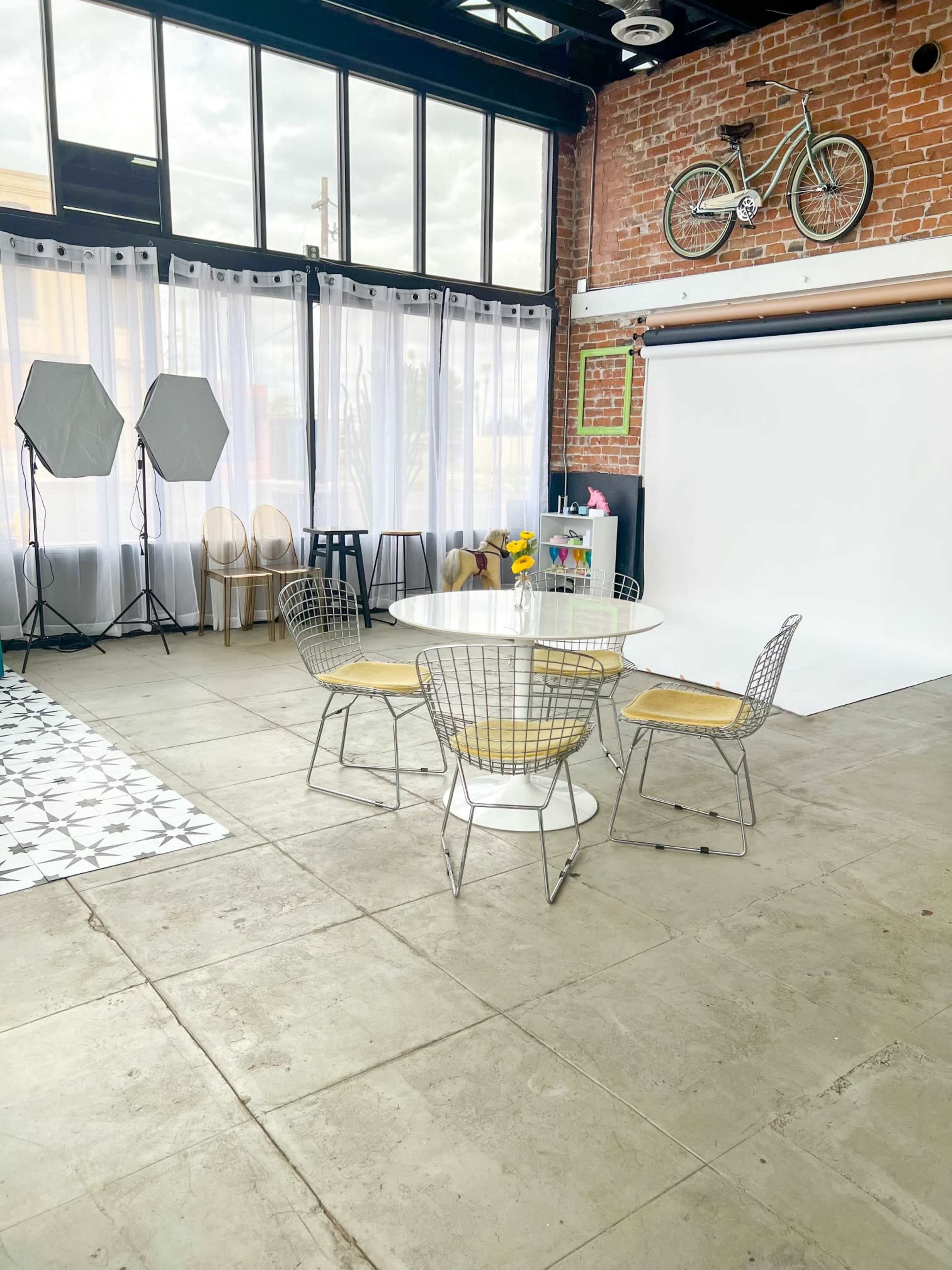 A modern studio space featuring a round table surrounded by wire chairs, large windows, and photography equipment in a brick-walled area.