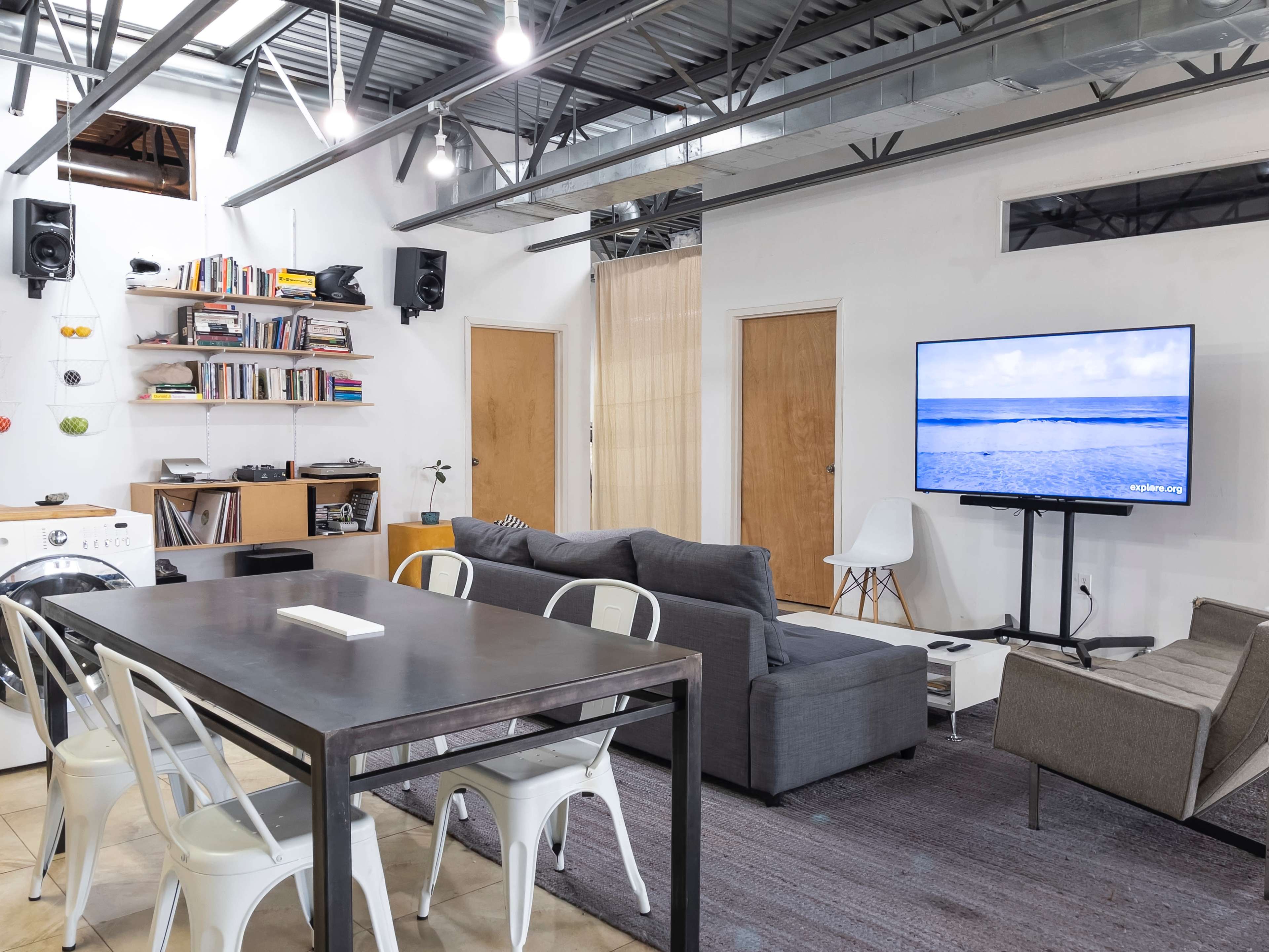 A modern, open-concept living space featuring a dining area with a metal table, a gray sectional sofa, a television on a stand, and bookshelves on the wall.