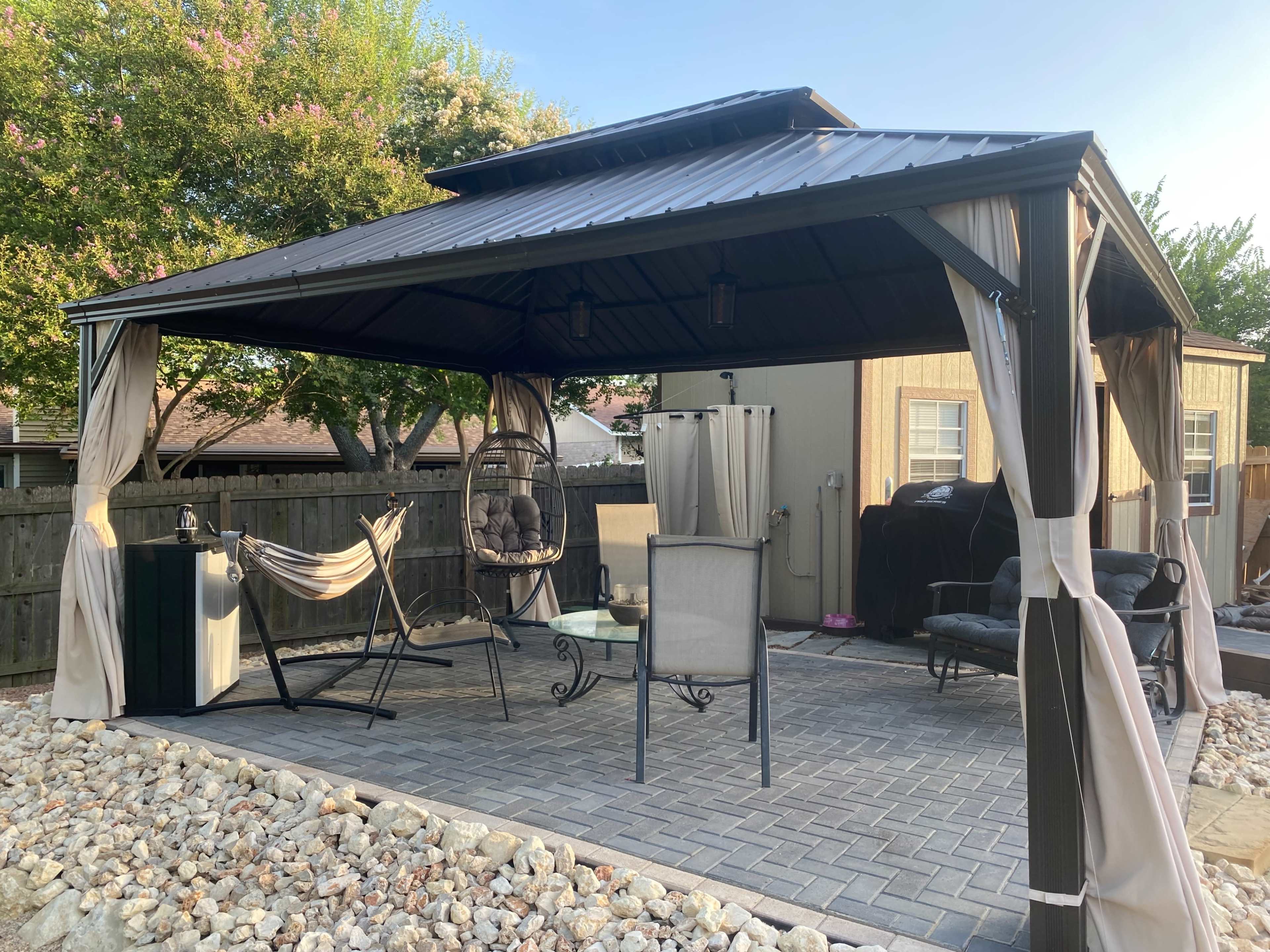 A metal gazebo with a high roof is furnished with several chairs and a hanging swing, surrounded by a stone patio and trees in the background.