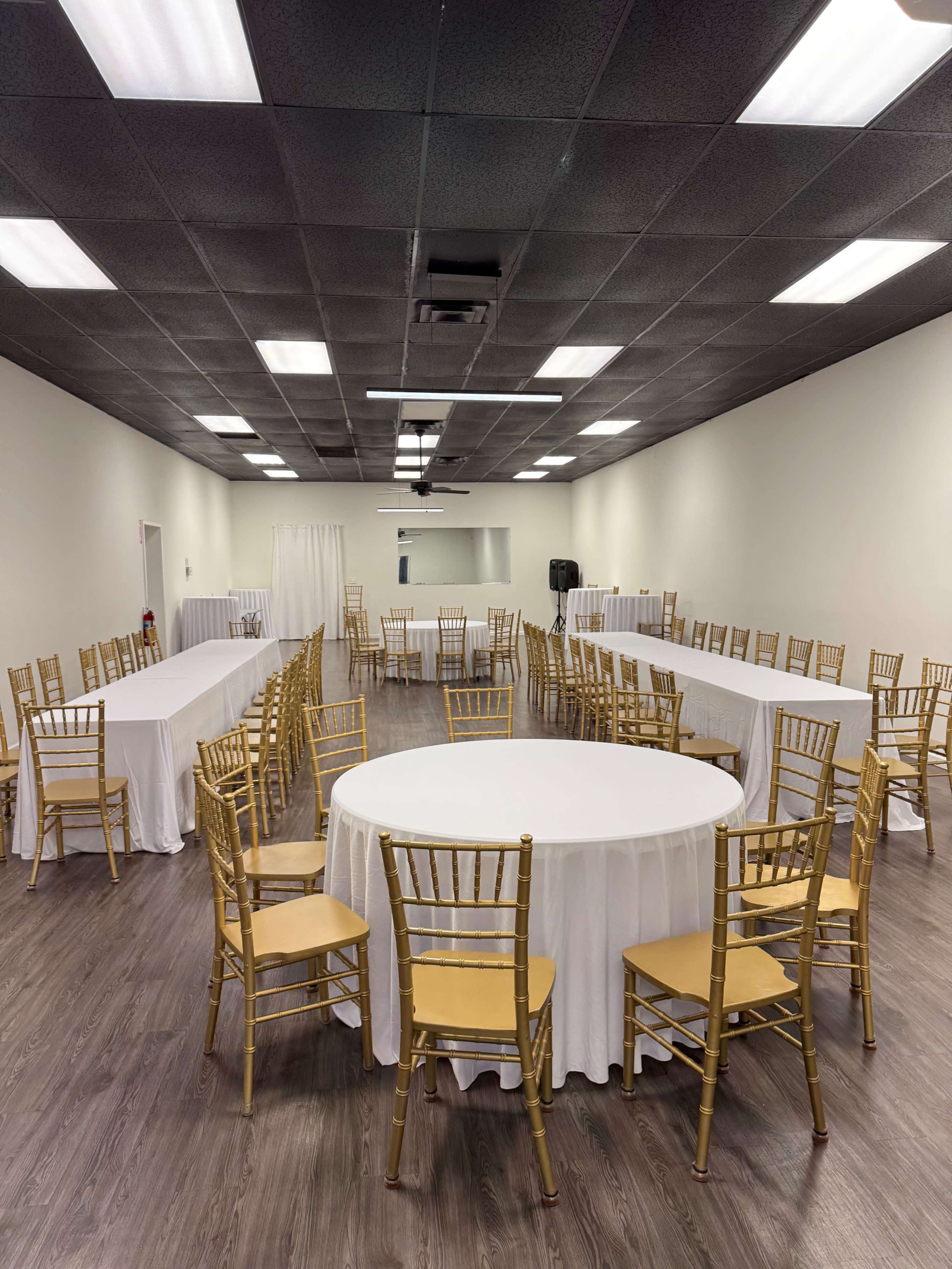 A spacious room featuring round tables covered with white tablecloths and surrounded by gold Chiavari chairs.