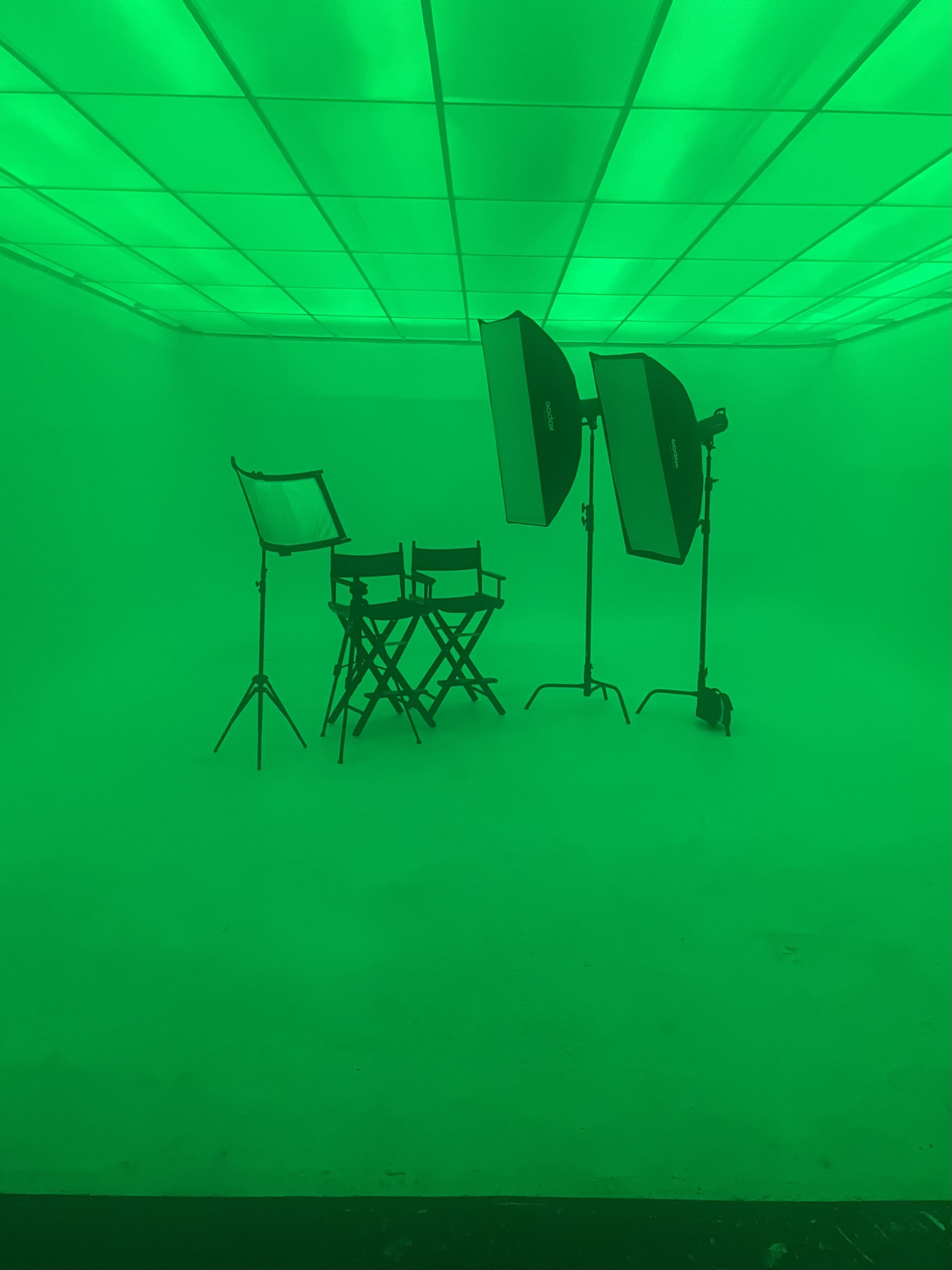 The image shows a dimly lit studio space with green walls and lighting, featuring two large softbox lights, a director's chair, and a standard chair arranged in front of them.