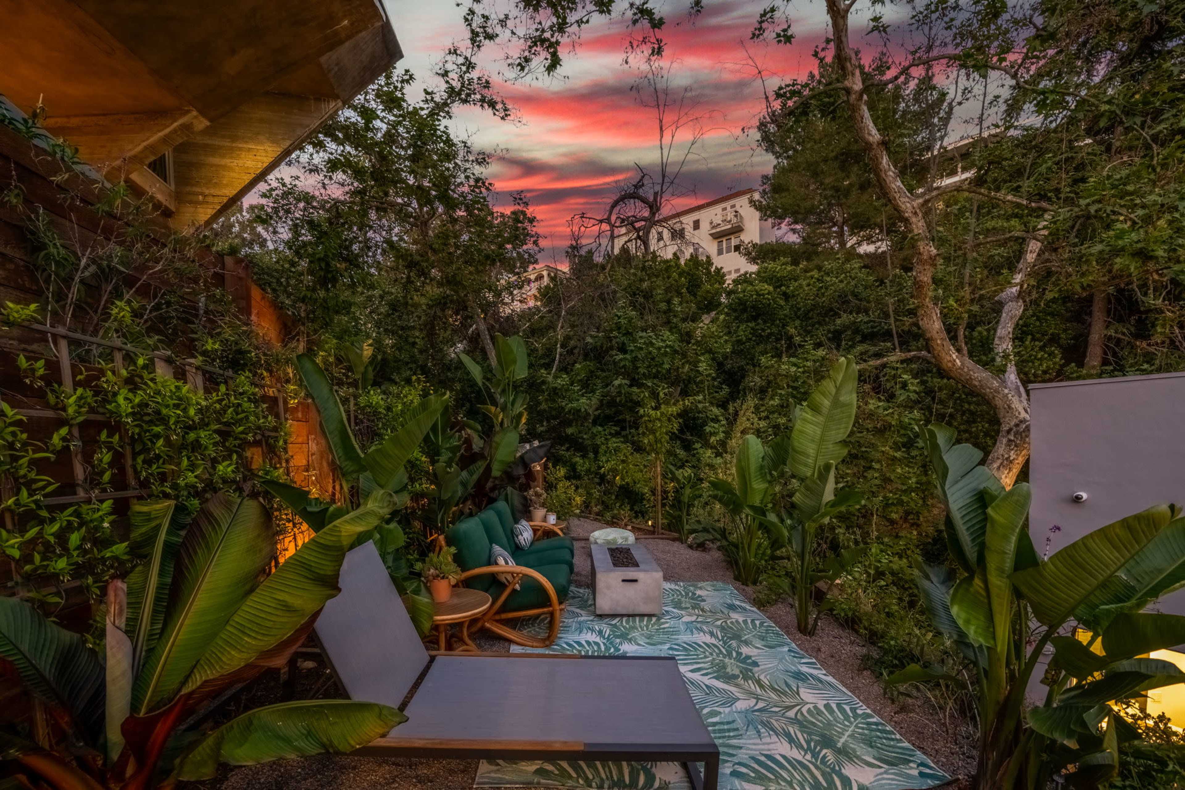 A cozy outdoor seating area is surrounded by lush greenery and colorful plants, set against a backdrop of a vibrant sunset sky.