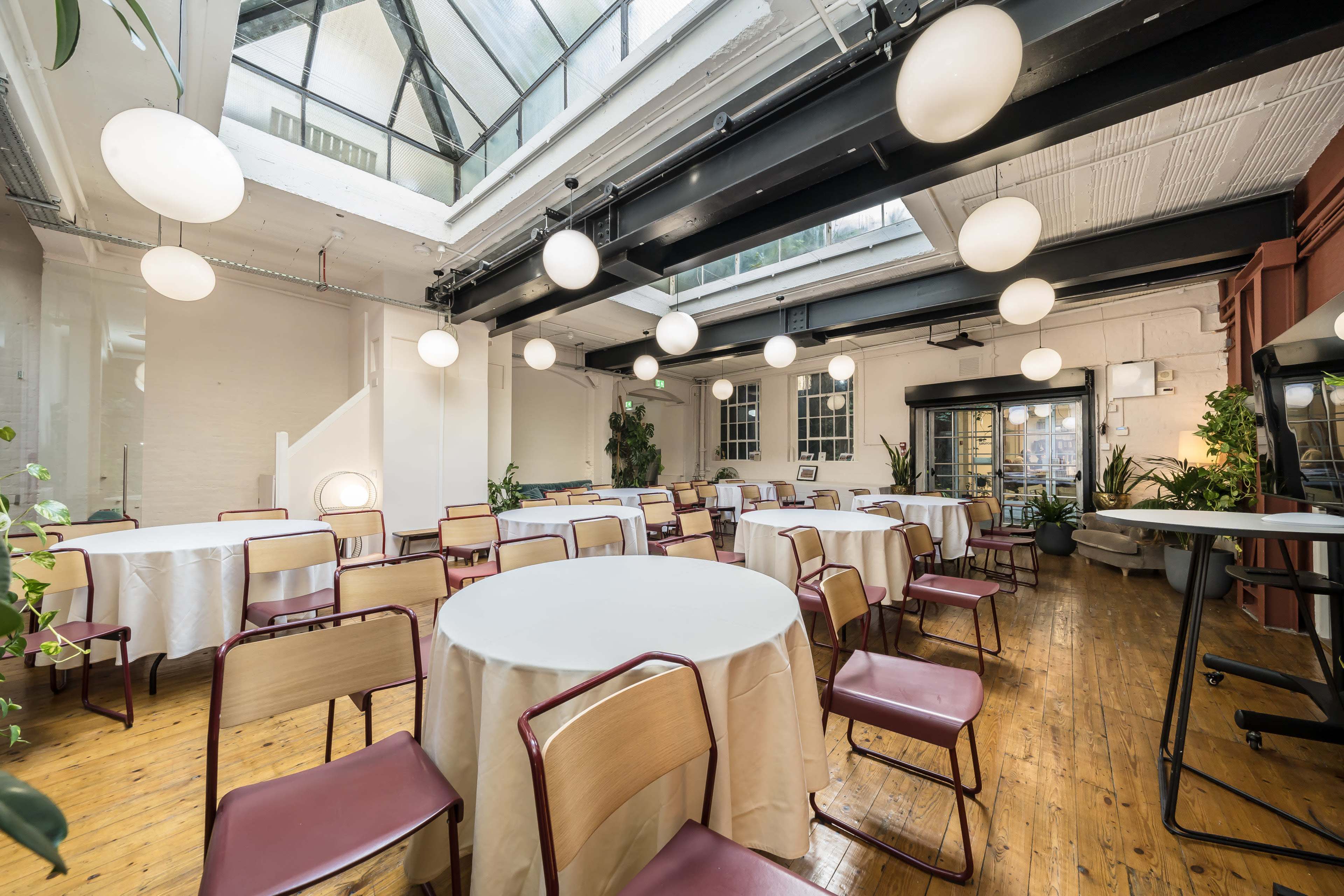 The image shows a spacious room with several round tables covered in white tablecloths, surrounded by chairs, and illuminated by large spherical lights hanging from the ceiling.