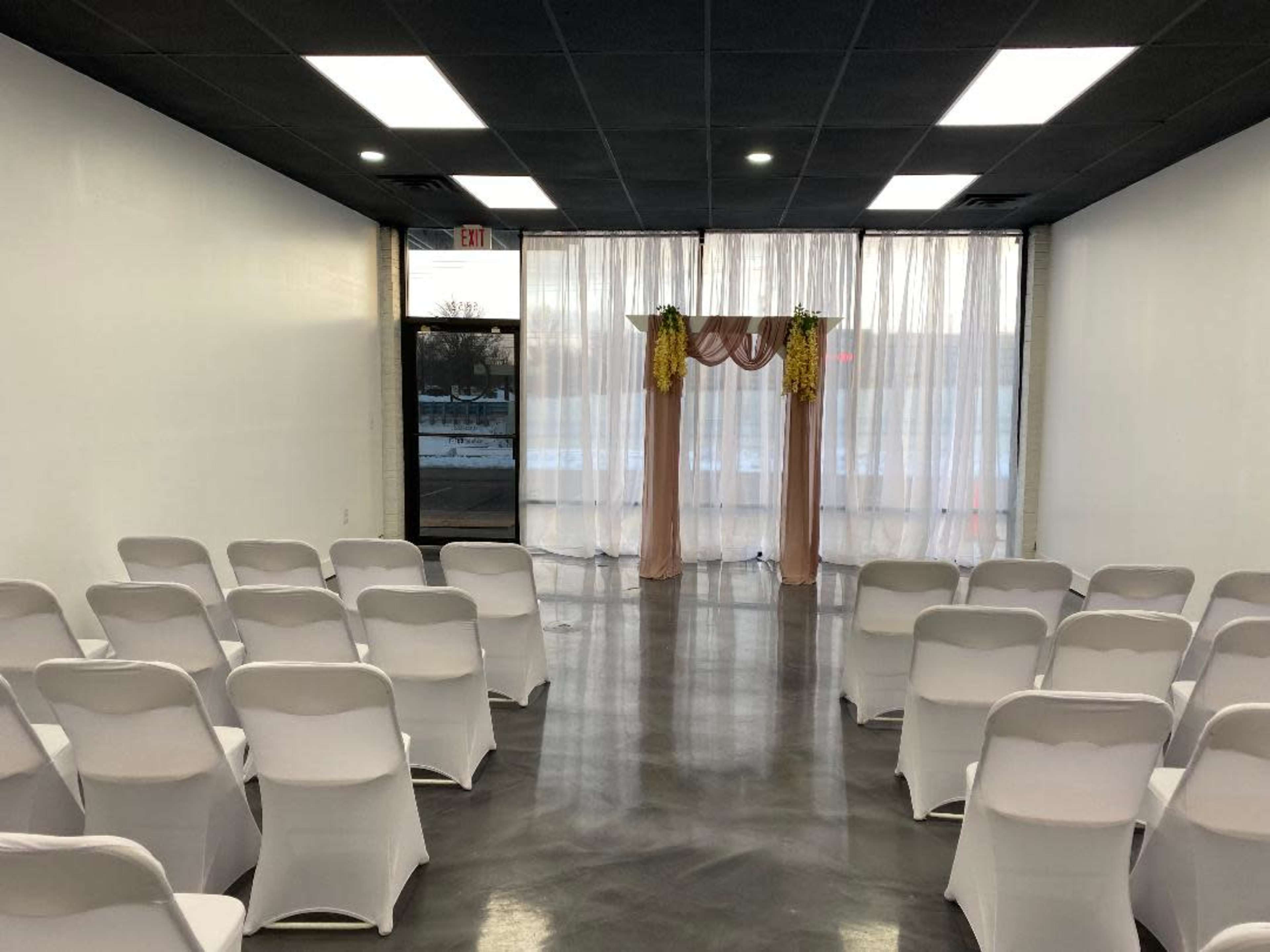 A well-lit event space with rows of white chairs facing a decorative arch draped with fabric at the front, near large windows.