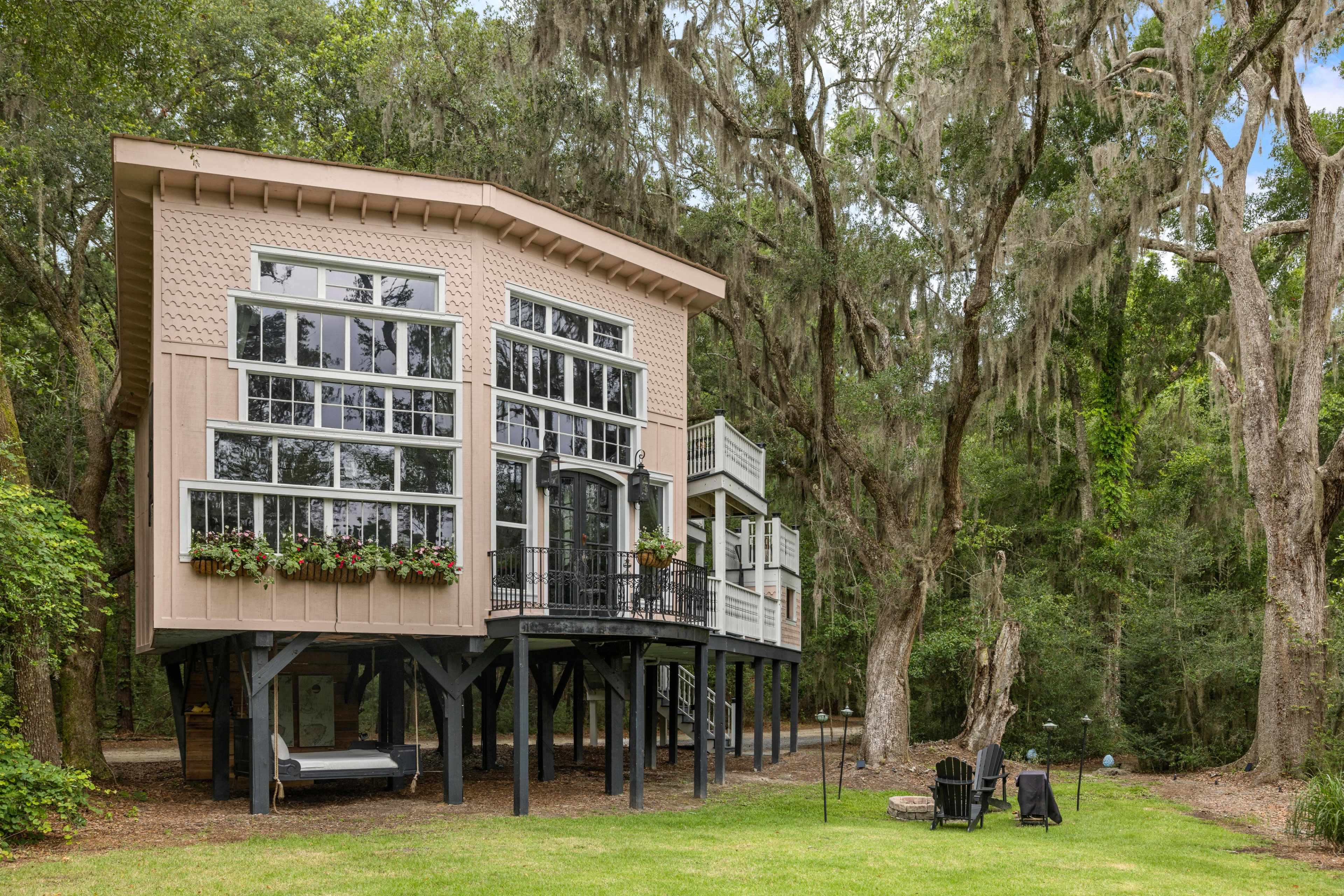 A two-story house on stilts surrounded by trees, with large windows and a patio deck, set in a grassy area.