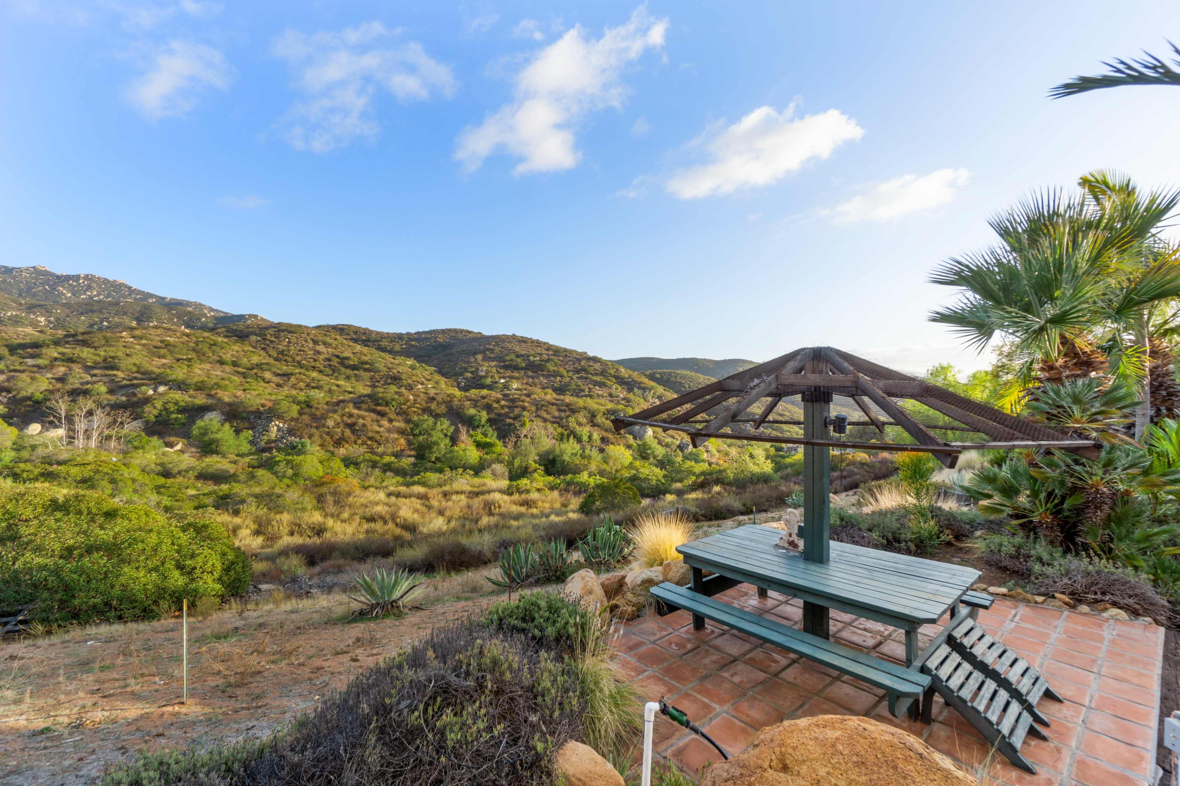 A wooden picnic table with a shaded gazebo overlooks a lush, hilly landscape under a blue sky.