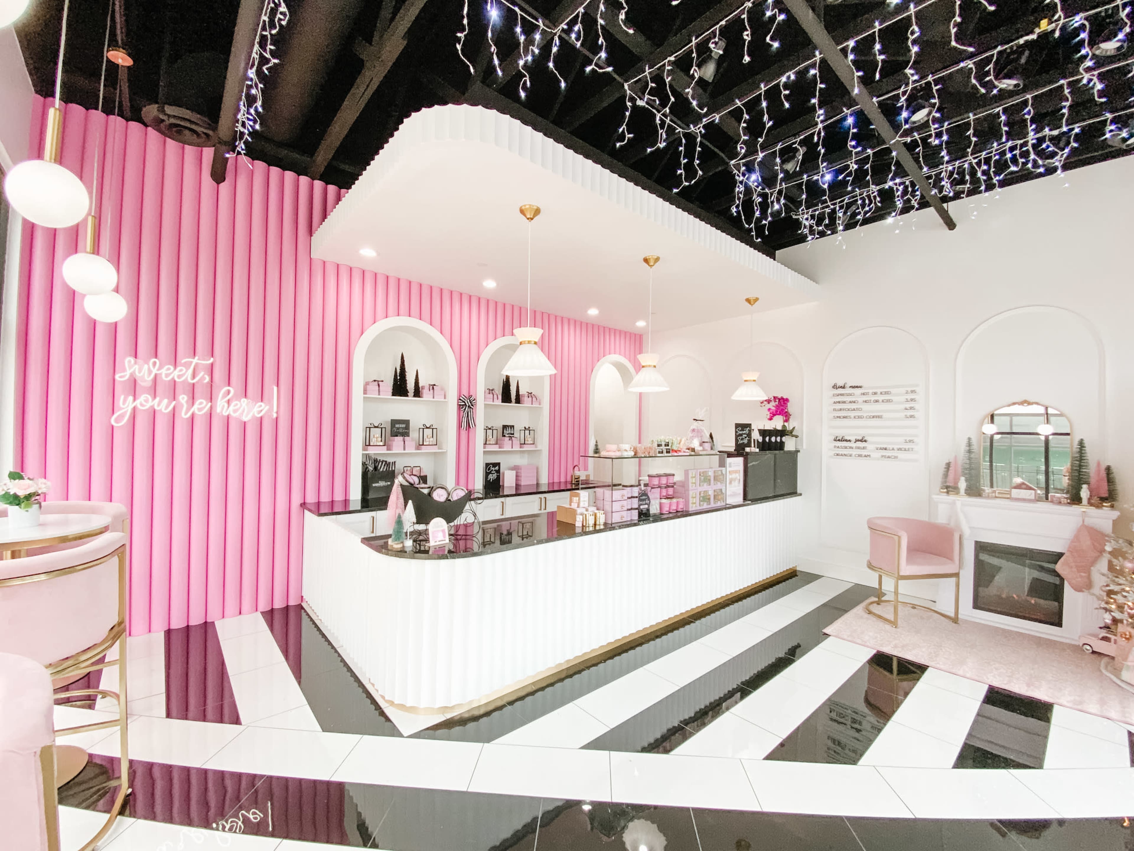A brightly lit beauty store features a sleek white and black counter, a pink accent wall with shelves, and decorative lighting overhead.