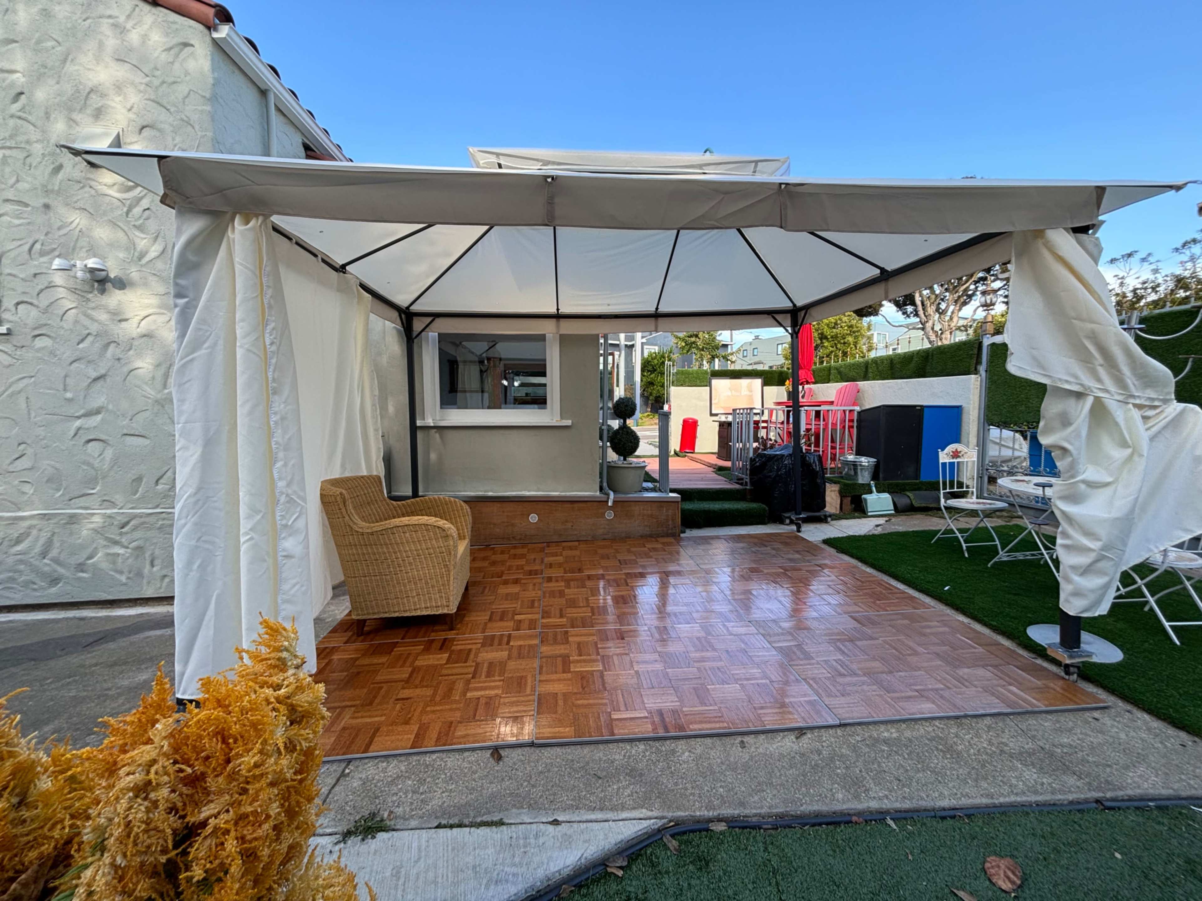 The image shows a covered patio area with a woven chair, wooden flooring, and a backdrop of walls and greenery.
