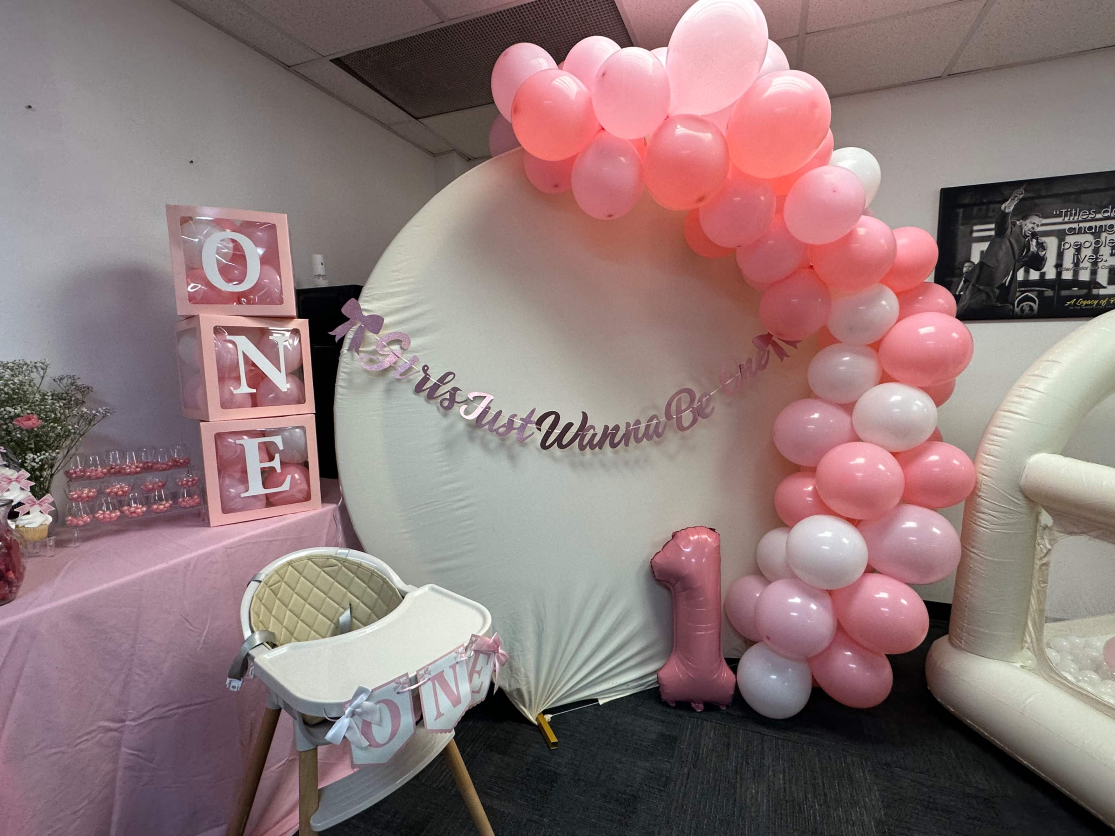 The image shows a decorated event space featuring a pink balloon arch, a large round backdrop with the phrase "#HersJustWannaBe1," and a table adorned with pink-themed decorations for a first birthday celebration.