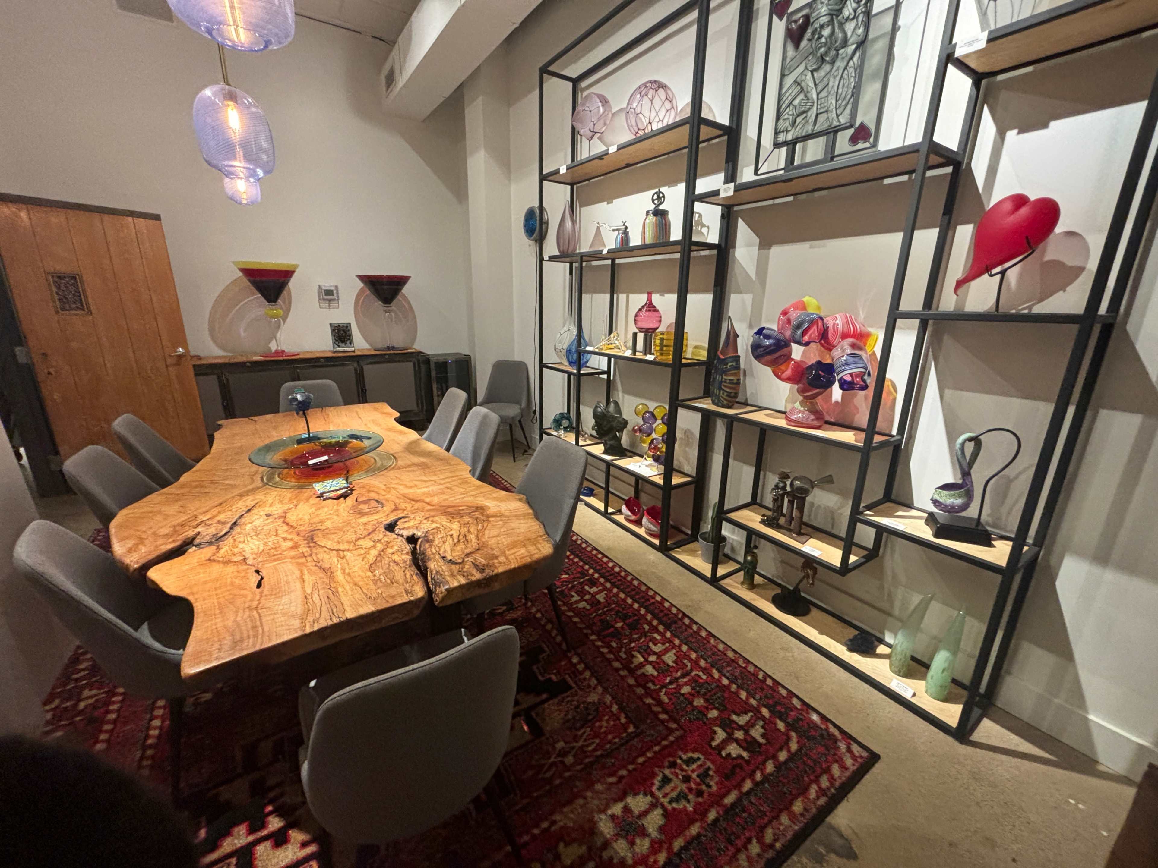 The image shows a modern dining area featuring a large wood table surrounded by gray chairs, with decorative shelves displaying various colorful glass art pieces and artifacts on the walls.