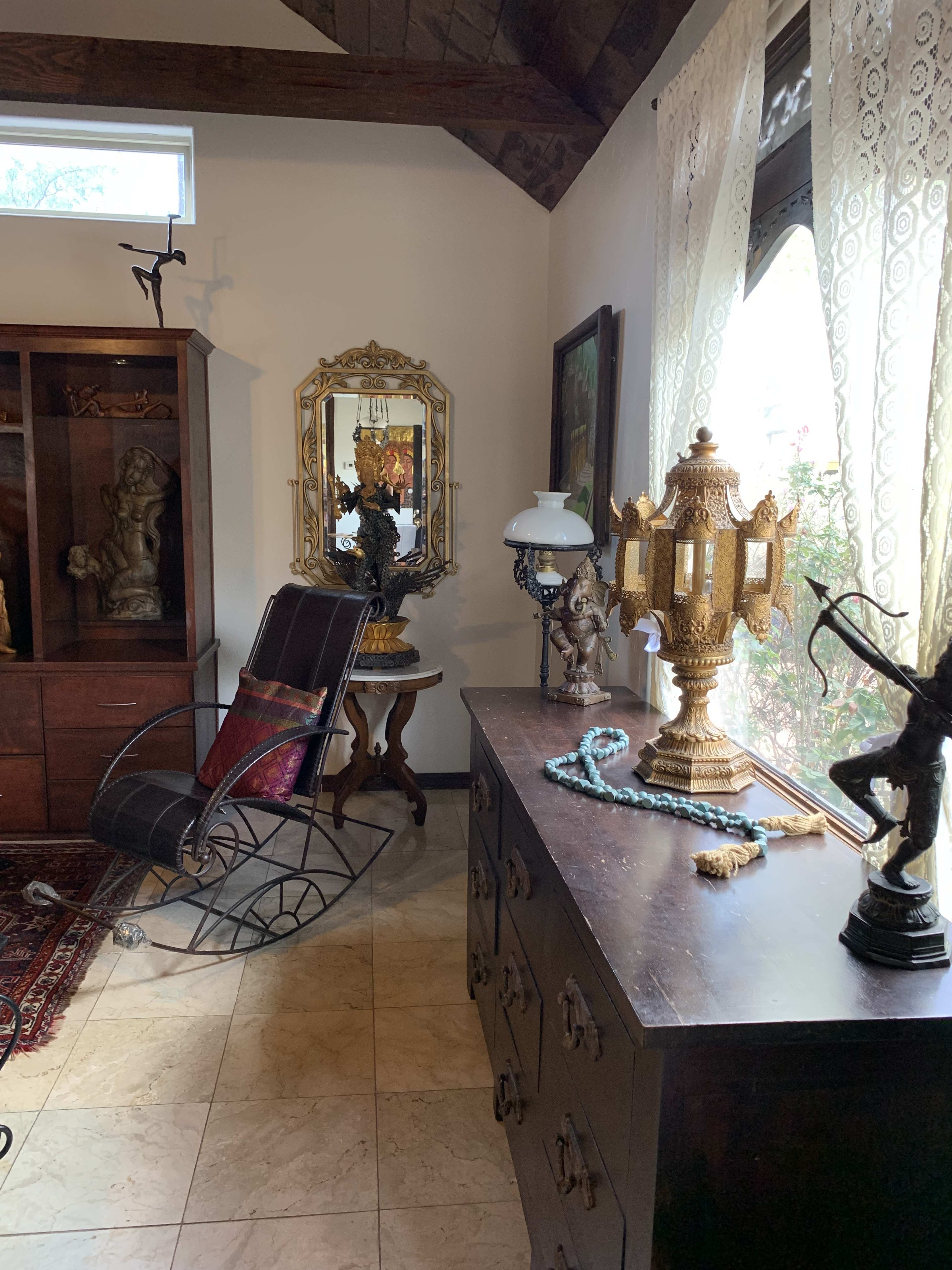 A room with a rocking chair, ornate lamps, and decorative sculptures, along with a wooden dresser and a window draped with lace curtains.