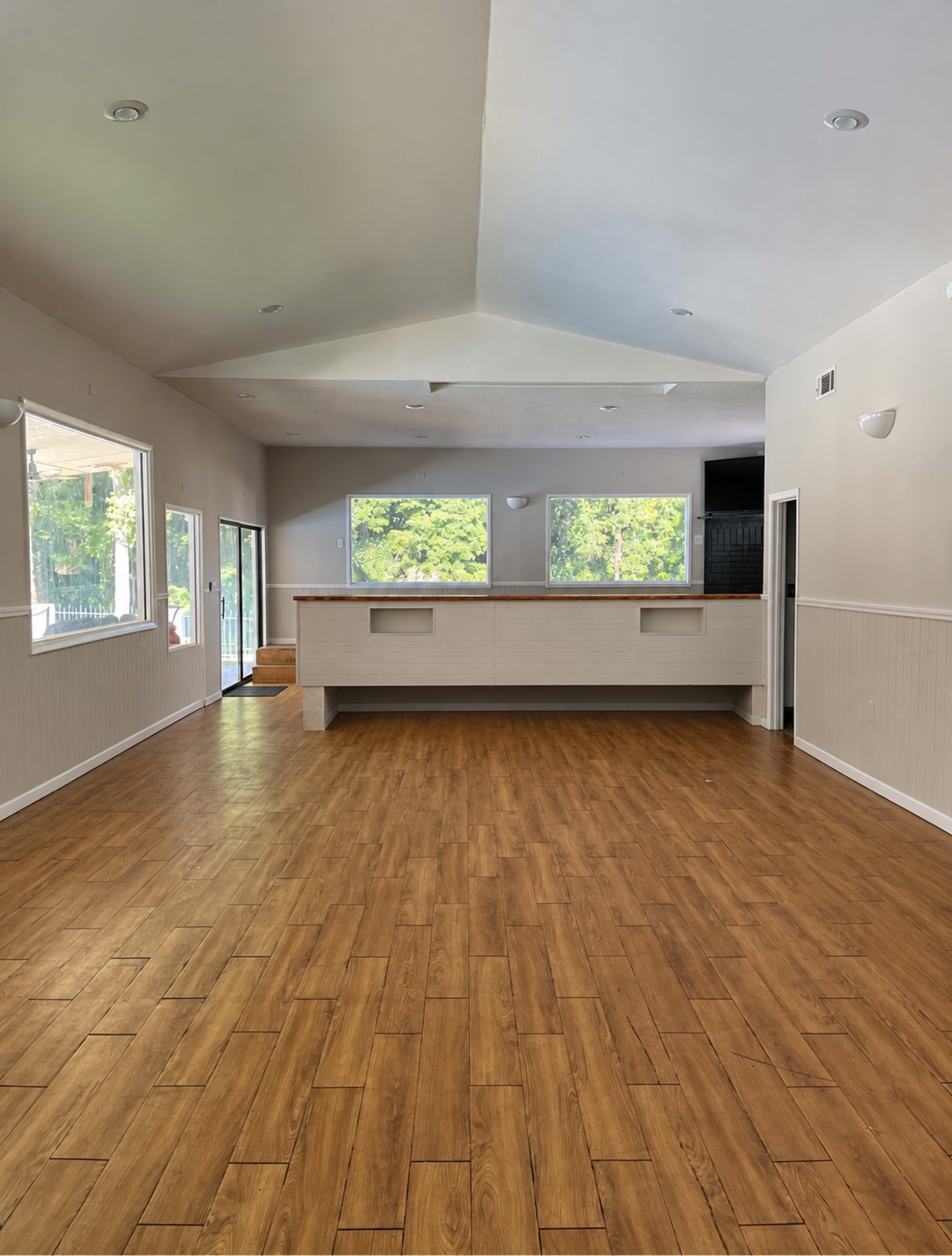 The image shows an empty room with a wooden floor, high ceiling, and a counter against one wall, flanked by large windows.
