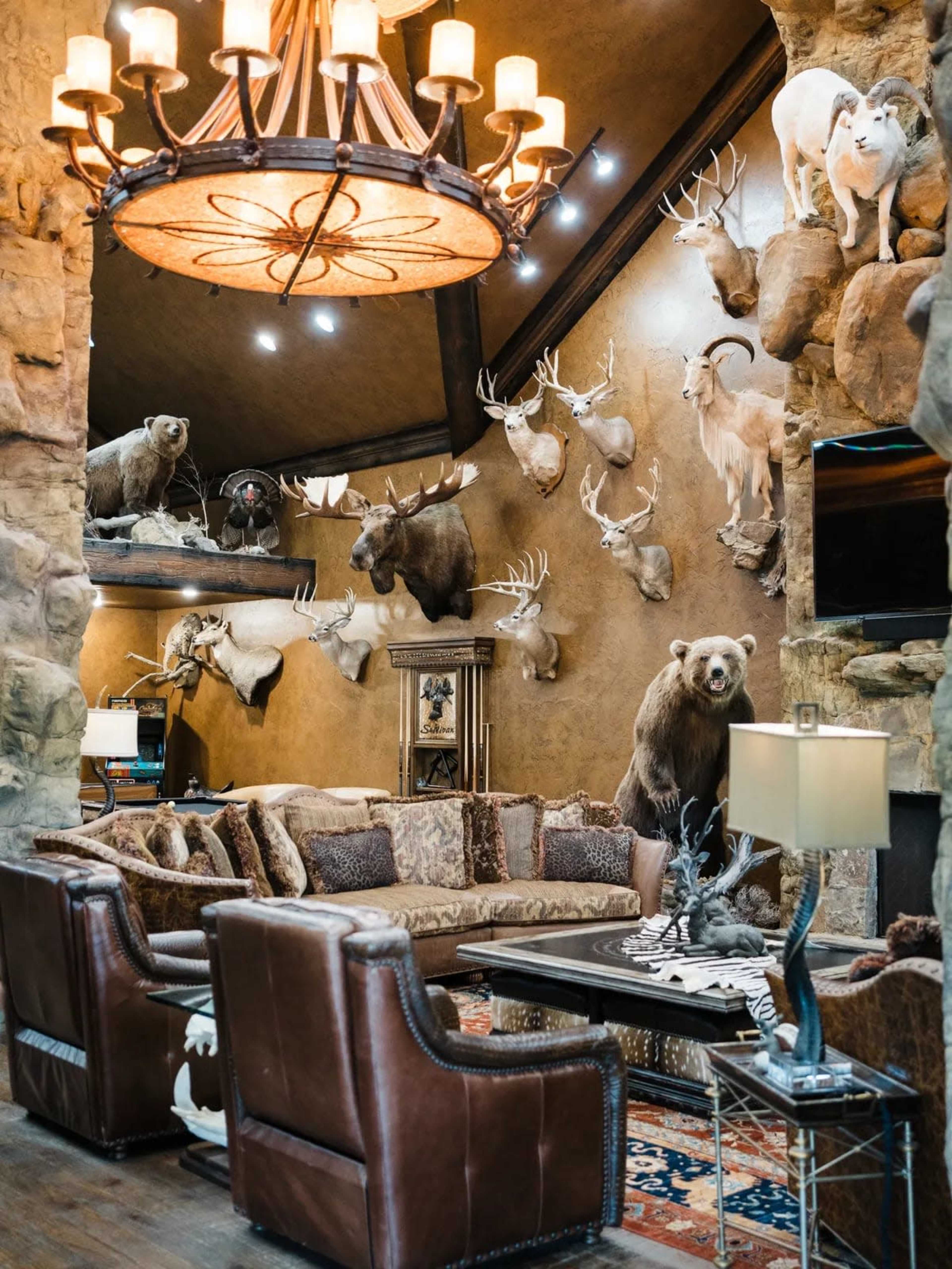 The image shows a rustic living room featuring a stone wall adorned with mounted animal trophies, a large chandelier overhead, and leather furniture arranged around a coffee table.