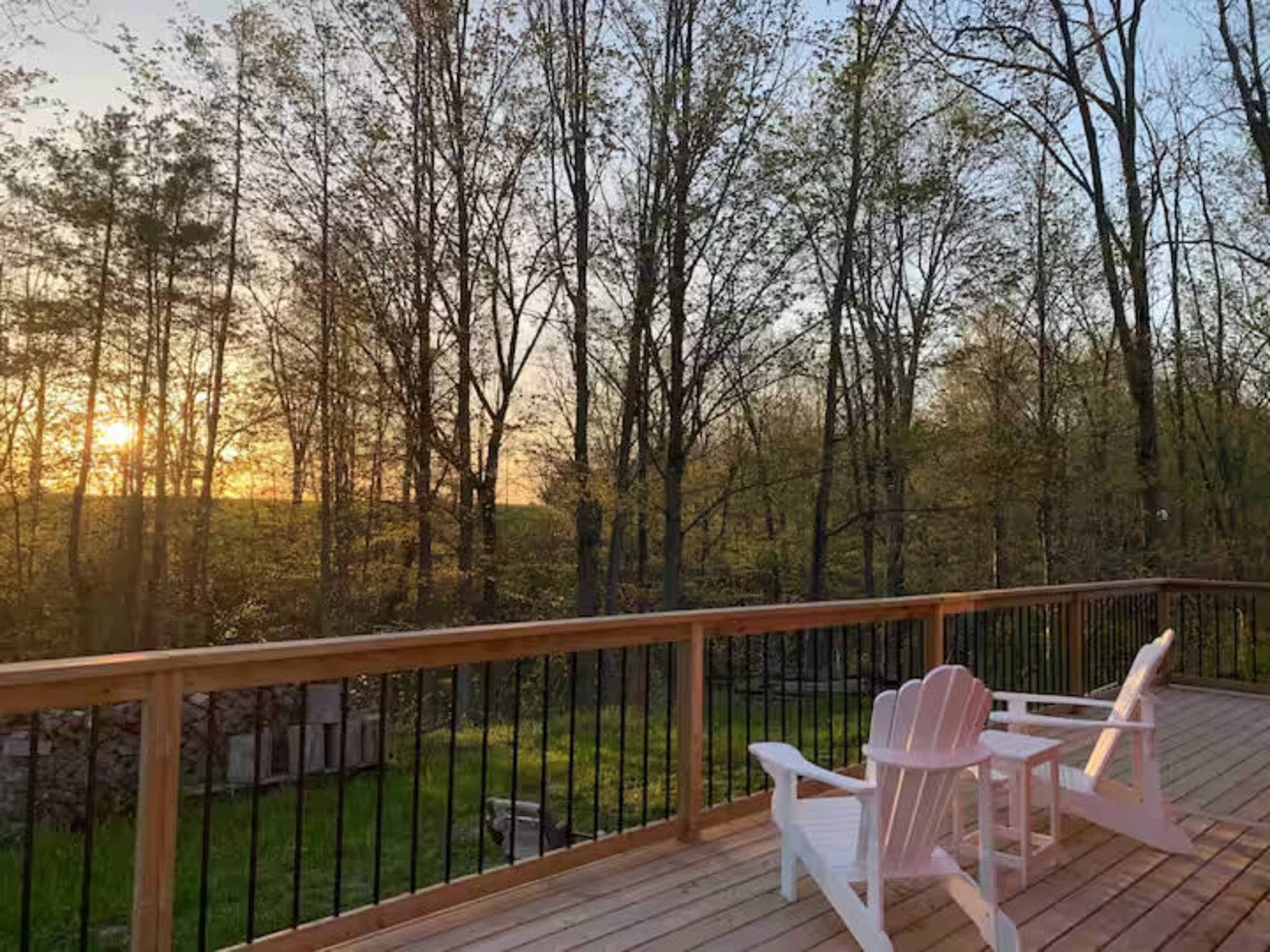 A wooden deck with two white Adirondack chairs overlooks a wooded area at sunset.