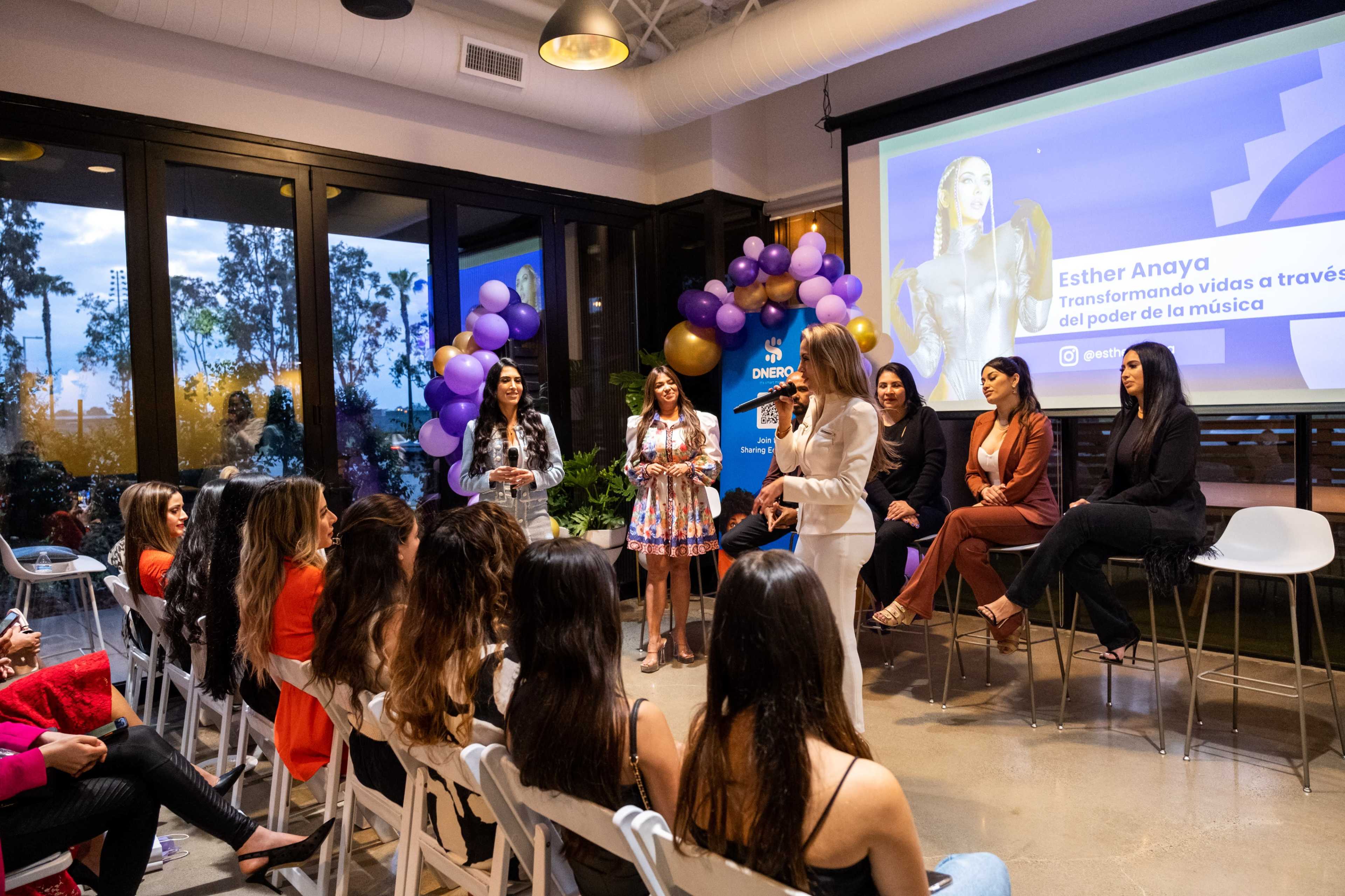 A group of women is gathered in a modern venue, with one woman speaking to the audience while others sit on a stage in the background, and decorative balloons are arranged nearby.