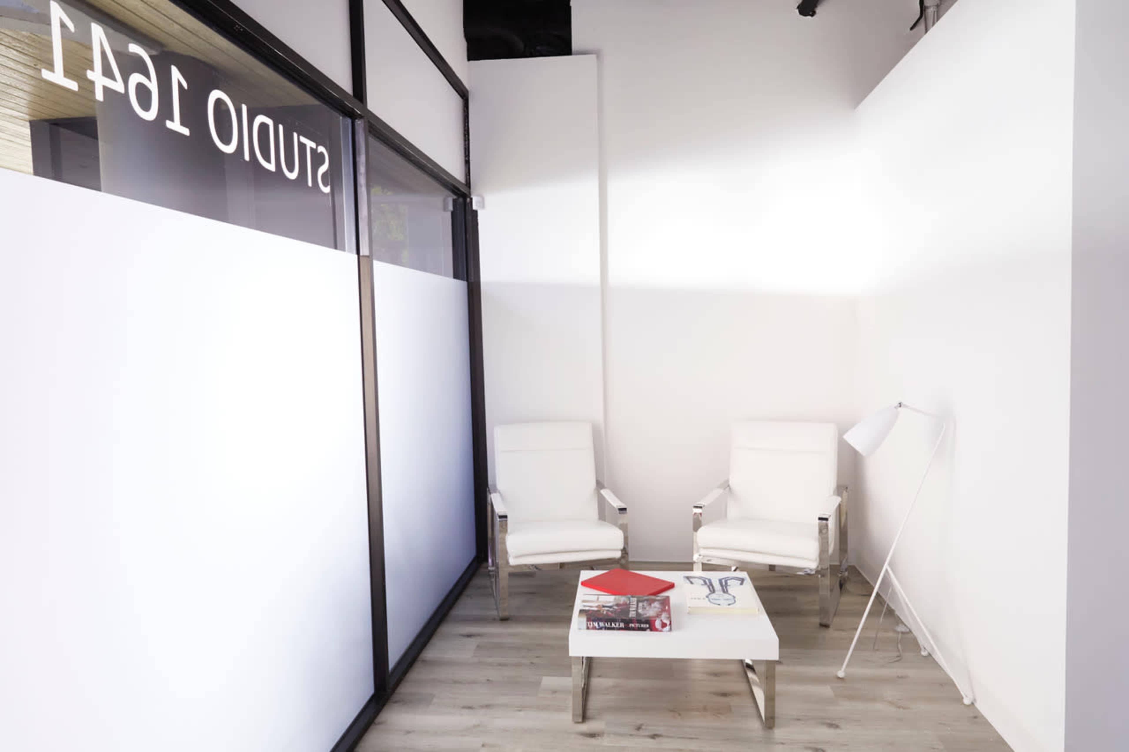 The image shows a minimalist waiting area with two white chairs, a small coffee table, and a floor lamp in a well-lit space.